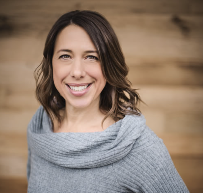 A smiling woman with shoulder-length brown hair, wearing a gray sweater, standing against a wooden background.