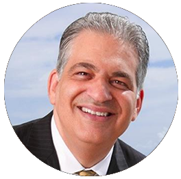 A smiling man with gray hair in a suit, outdoors with a partly cloudy sky background.