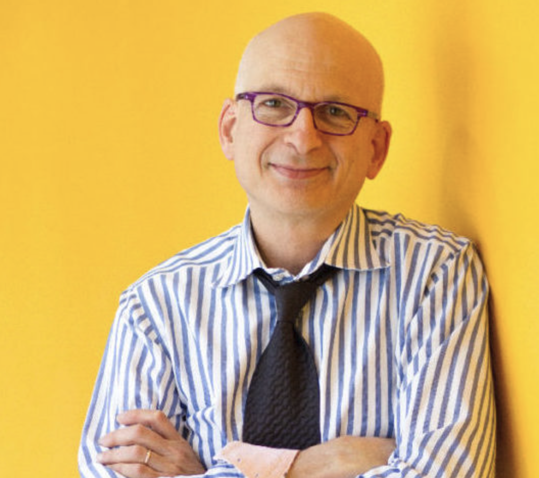 Bald man wearing glasses, a striped shirt, and a black tie, standing with arms crossed against a yellow background, smiling.