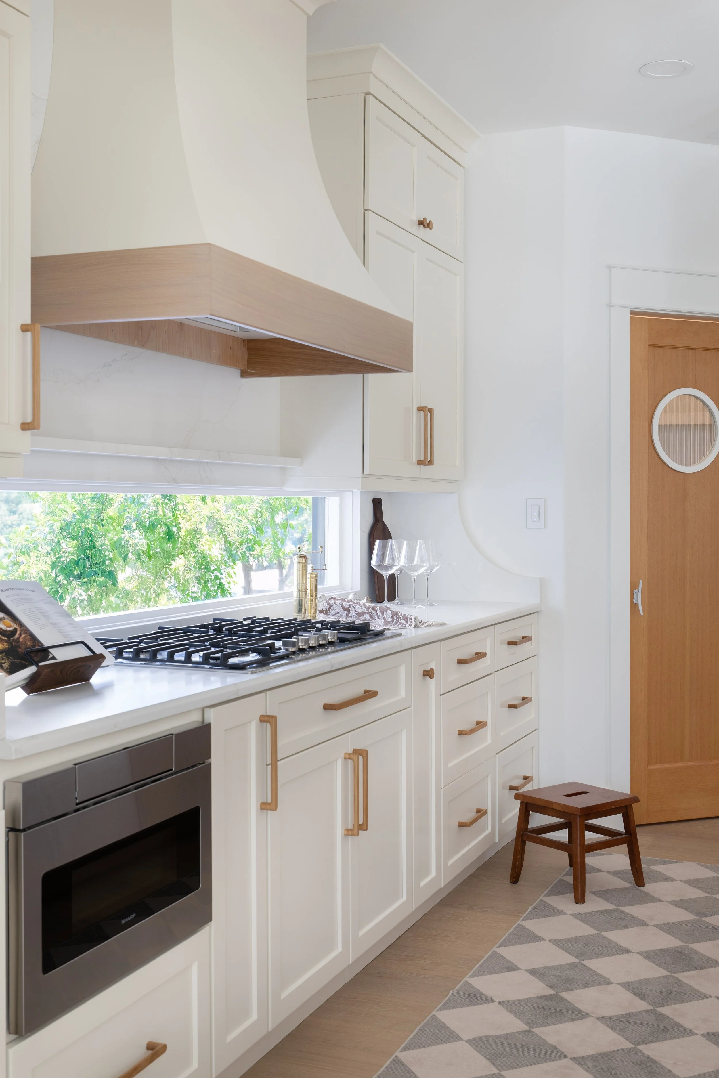 White kitchen with wooden accents, stainless steel oven, gas cooktop, white countertop, window with green trees outside, small wooden stool, and patterned gray and white rug.