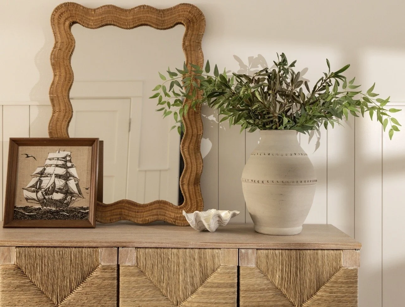 Decorative wooden sideboard with a framed nautical picture, a wavy-edged mirror, a large ceramic vase with green leafy plant, and a white shell-shaped dish.