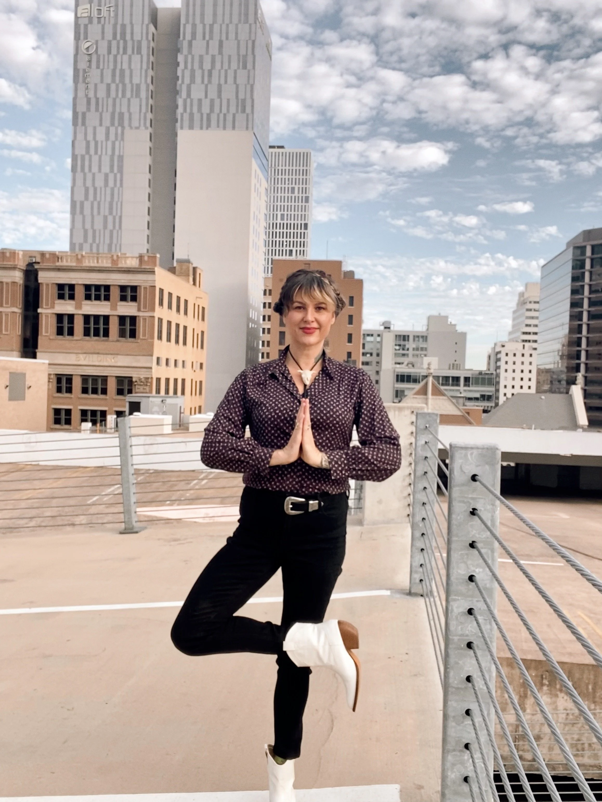 A woman practicing yoga outdoors on a rooftop with a city skyline in the background, standing in a tree pose with hands in a prayer position at her chest.