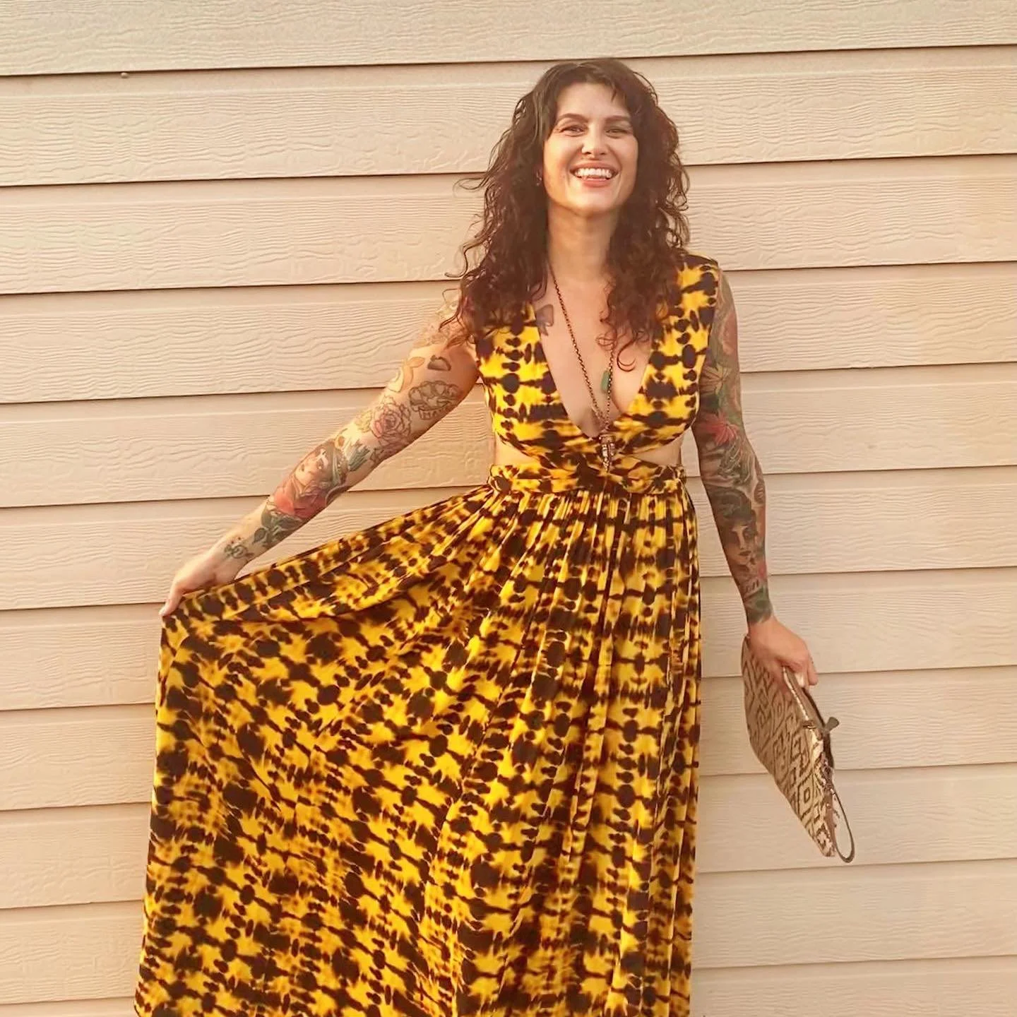 Woman with curly hair wearing a yellow and black animal print dress, standing in front of a beige wooden wall, smiling, holding the hem of her dress and a purse.