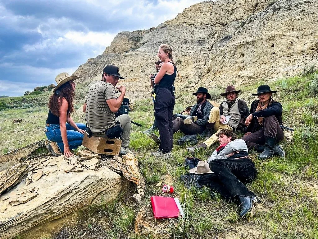Group of people on a film set outdoors, with rocky terrain and cliffs in the background. Some actors are in costume, others are crew members, and they are sitting or kneeling on the grass.