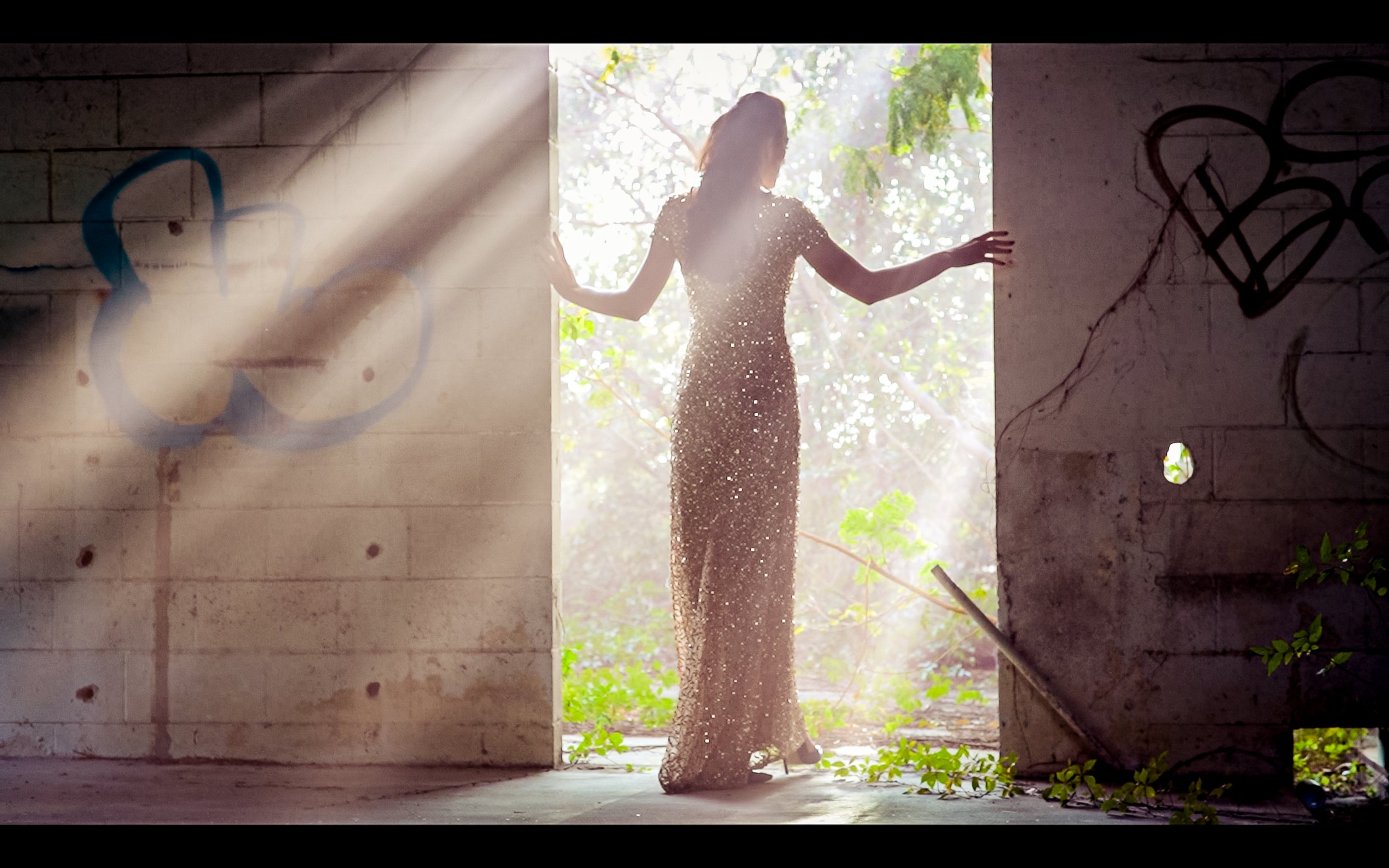 A woman in a sparkly dress with her arms outstretched stands in an open doorway, with sunlight streaming in and outdoor greenery visible through the opening.