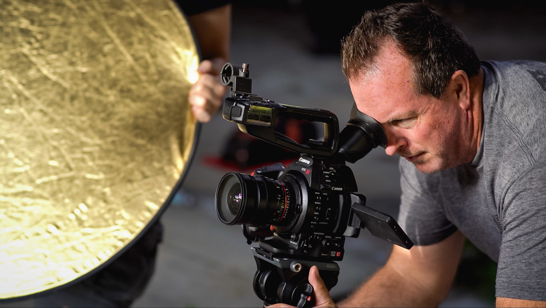A man looking through a professional video camera lens and filming a subject with a large gold reflector in the foreground.