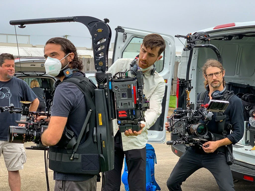 Three men working with professional camera equipment near an open van on a cloudy day.