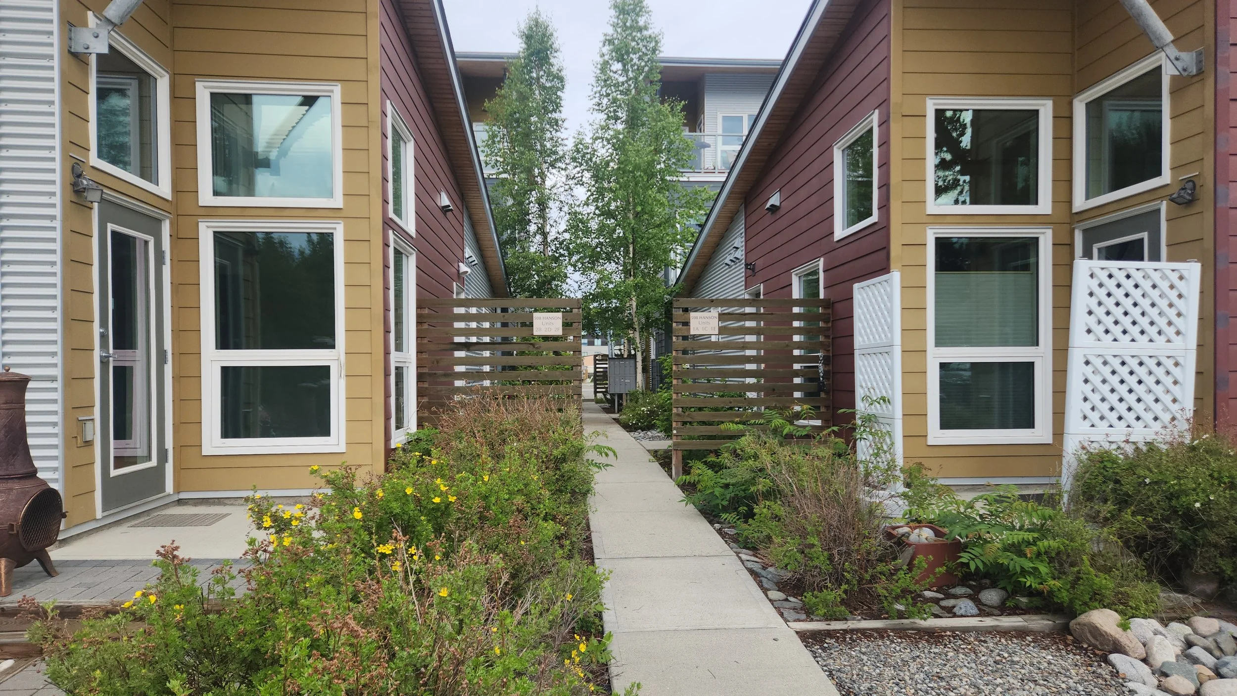 View of modern multi-story apartment buildings with colorful siding, large windows, and a shared walkway with landscaped plants and trees in a residential complex.