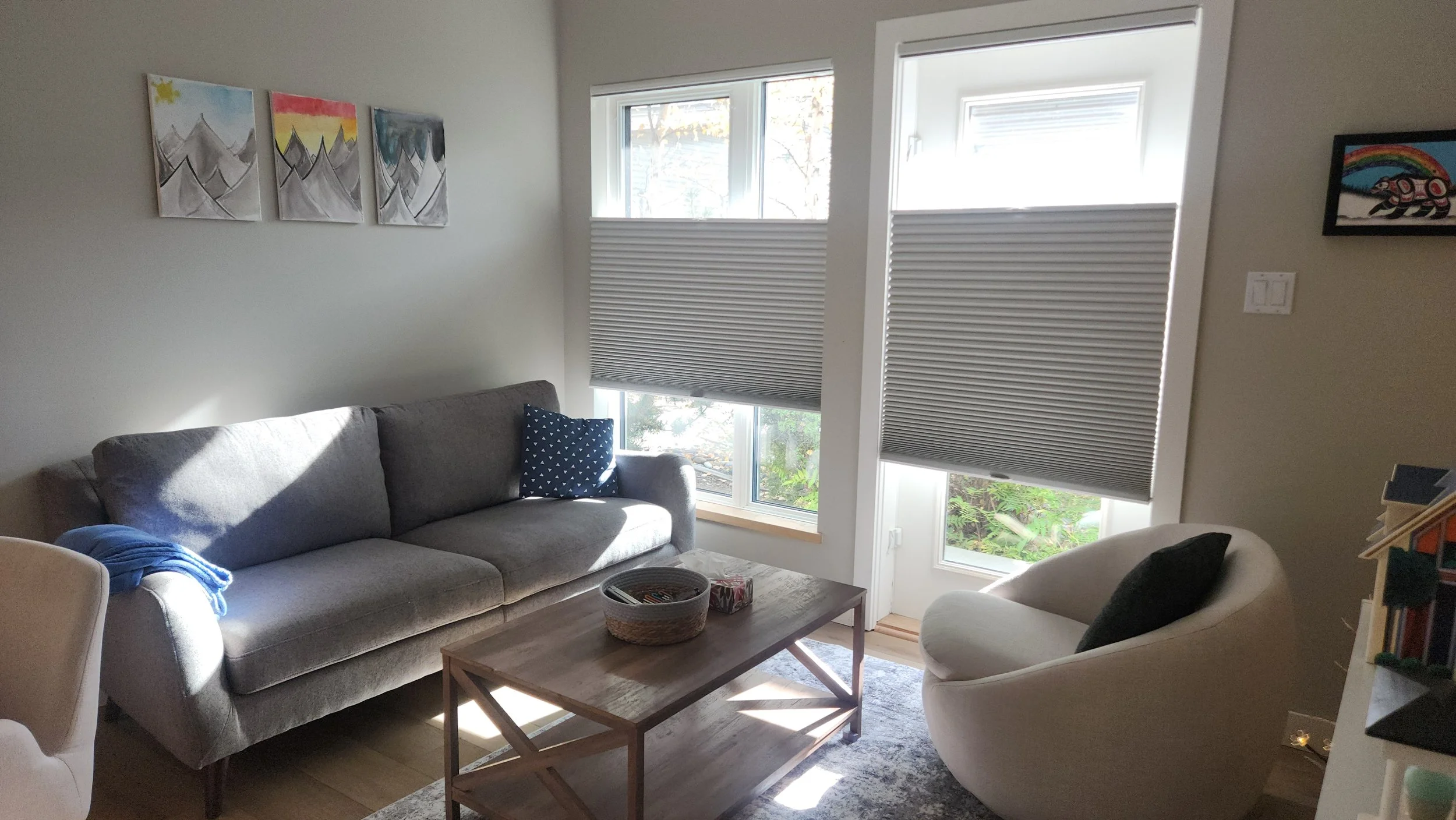 A cozy living room with a light gray sofa, a white armchair, and a wooden coffee table. Three artwork pieces of mountain scenes hang on the wall, and large windows with partly closed blinds let in sunlight.
