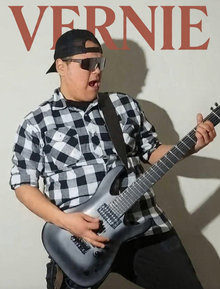 Young person wearing sunglasses, a backwards baseball cap, and a black-and-white checkered shirt, playing a gray electric guitar with an expression of enjoyment against a plain wall with the word 'VERNIE' in red letters.