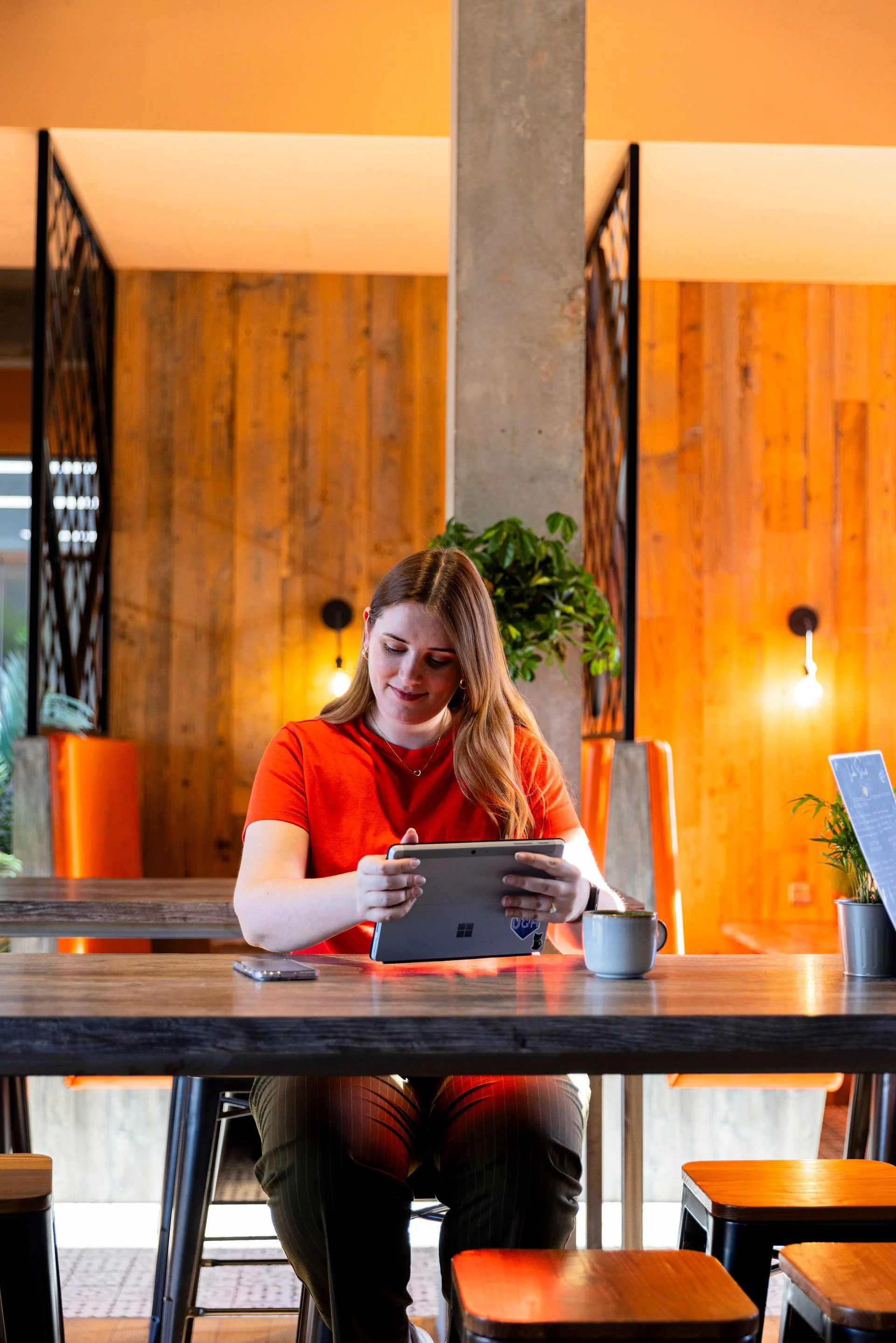 A woman in a red shirt sitting at a wooden table in a modern cafe, holding a tablet, with a cup, a smartphone, and a small plant on the table.