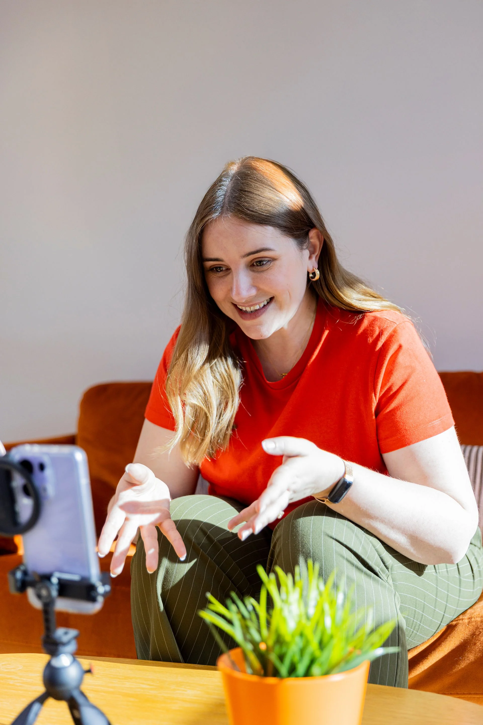 A woman with long blonde hair, wearing a red shirt and green pants, is smiling and talking while recording herself on a smartphone during a video call or recording. She is sitting on a couch with a plant in an orange pot and a plain background.
