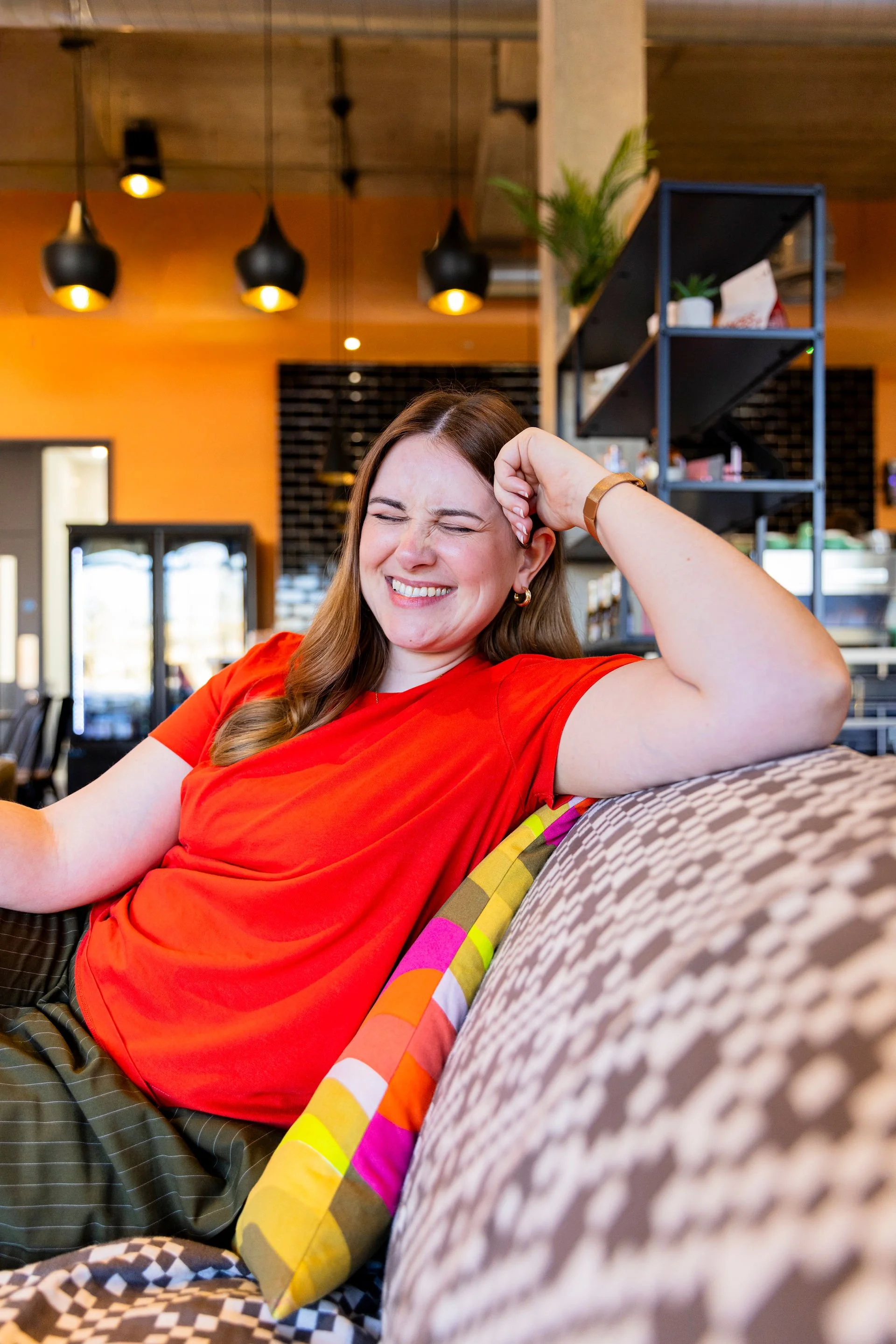 A woman with brown hair wearing a red shirt and striped pants sitting on a couch, smiling with eyes closed and stretching her arm while in a modern indoor space.
