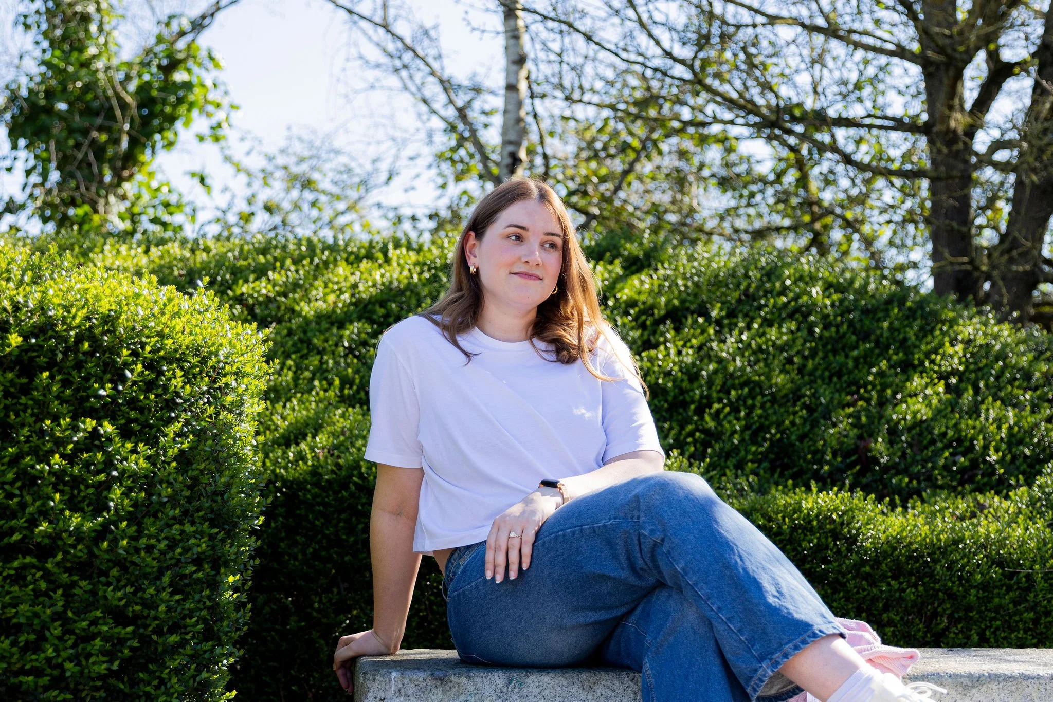 A young woman with brown hair sitting on a stone ledge outdoors, surrounded by greenery and trees, wearing a white t-shirt and blue jeans
