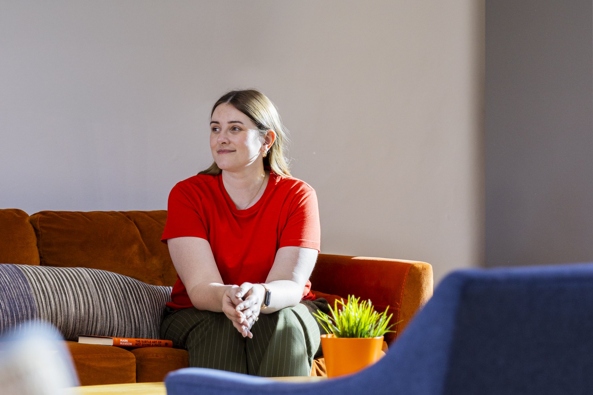 A woman with light brown hair, wearing a red shirt, sitting on a brown couch with striped and solid cushions, smiling and looking to her left. There is a small green potted plant and a red book on the table in front of her.