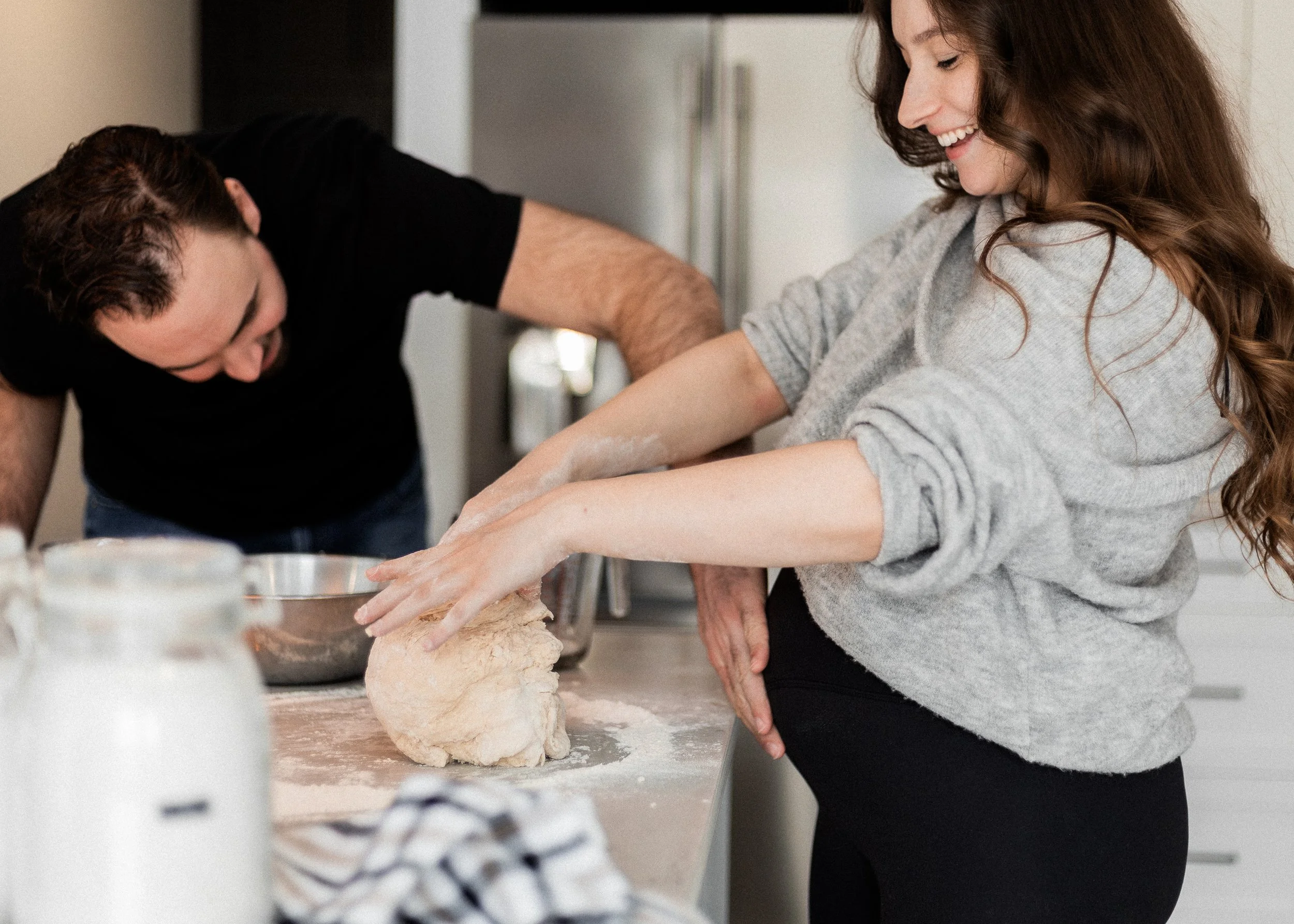 A woman with long brown hair and a light gray sweater smiling as she kneads dough on a kitchen counter. A man with short dark hair and a black shirt leans over her, possibly helping or participating in baking. The kitchen has a modern, minimal design.