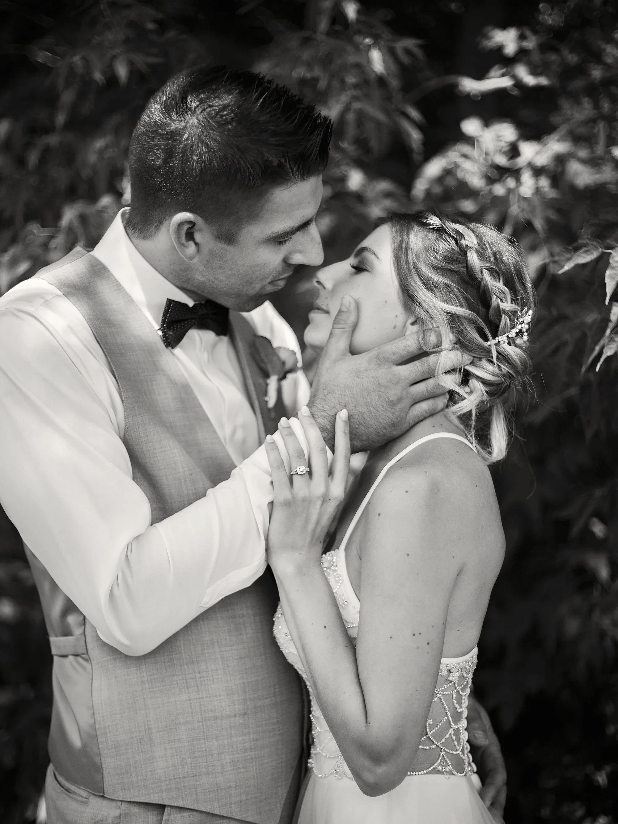 A black and white photograph of a couple in wedding attire, standing close and touching foreheads with eyes closed, amidst blurred foliage background.