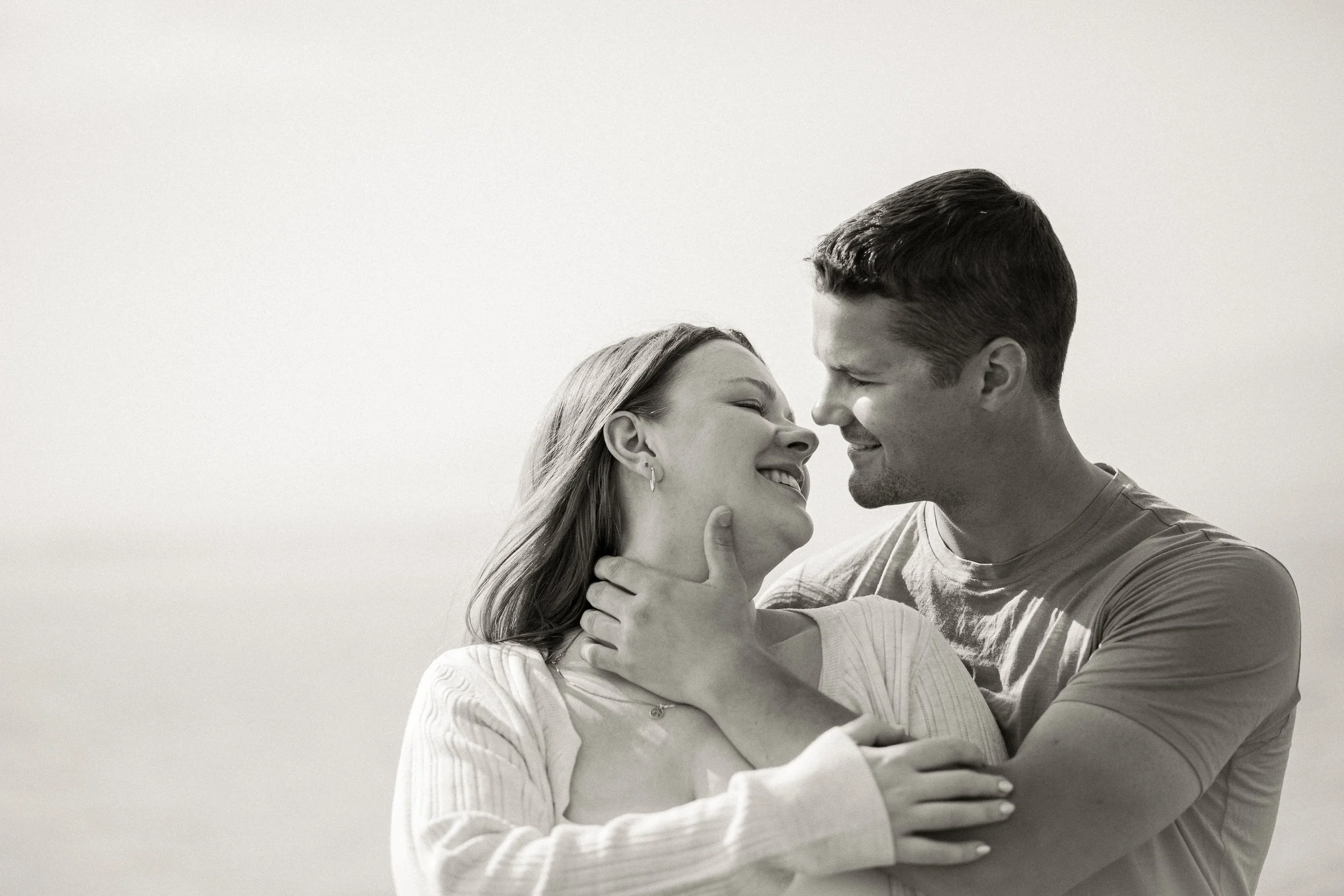 A black-and-white photo of a smiling couple looking at each other closely, with the woman touching the man's face and the man holding the woman gently.