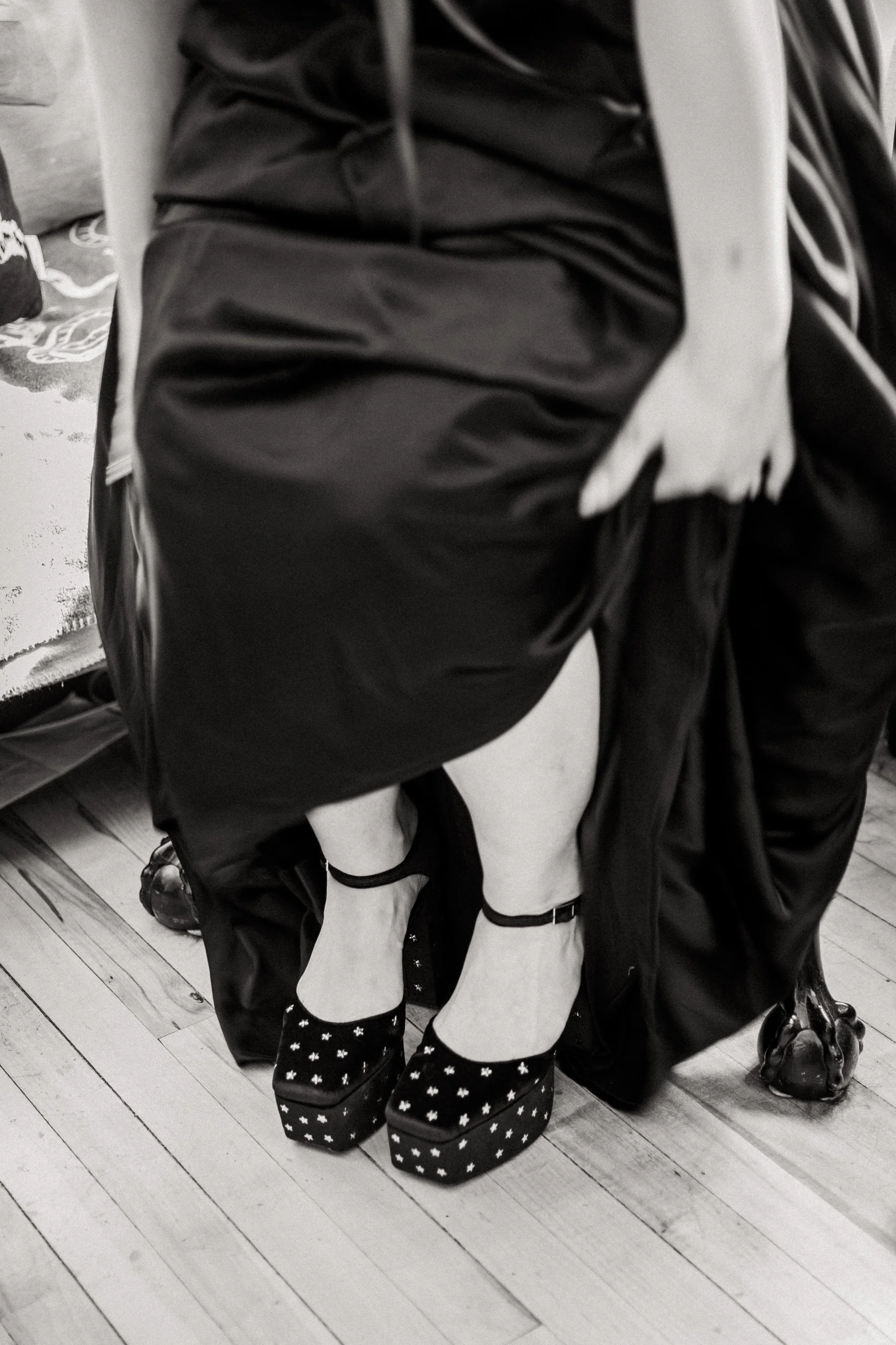 Close-up of a person sitting on a chair with their legs crossed, wearing black boots with white star patterns, white stockings, and black socks. The floor is wooden.