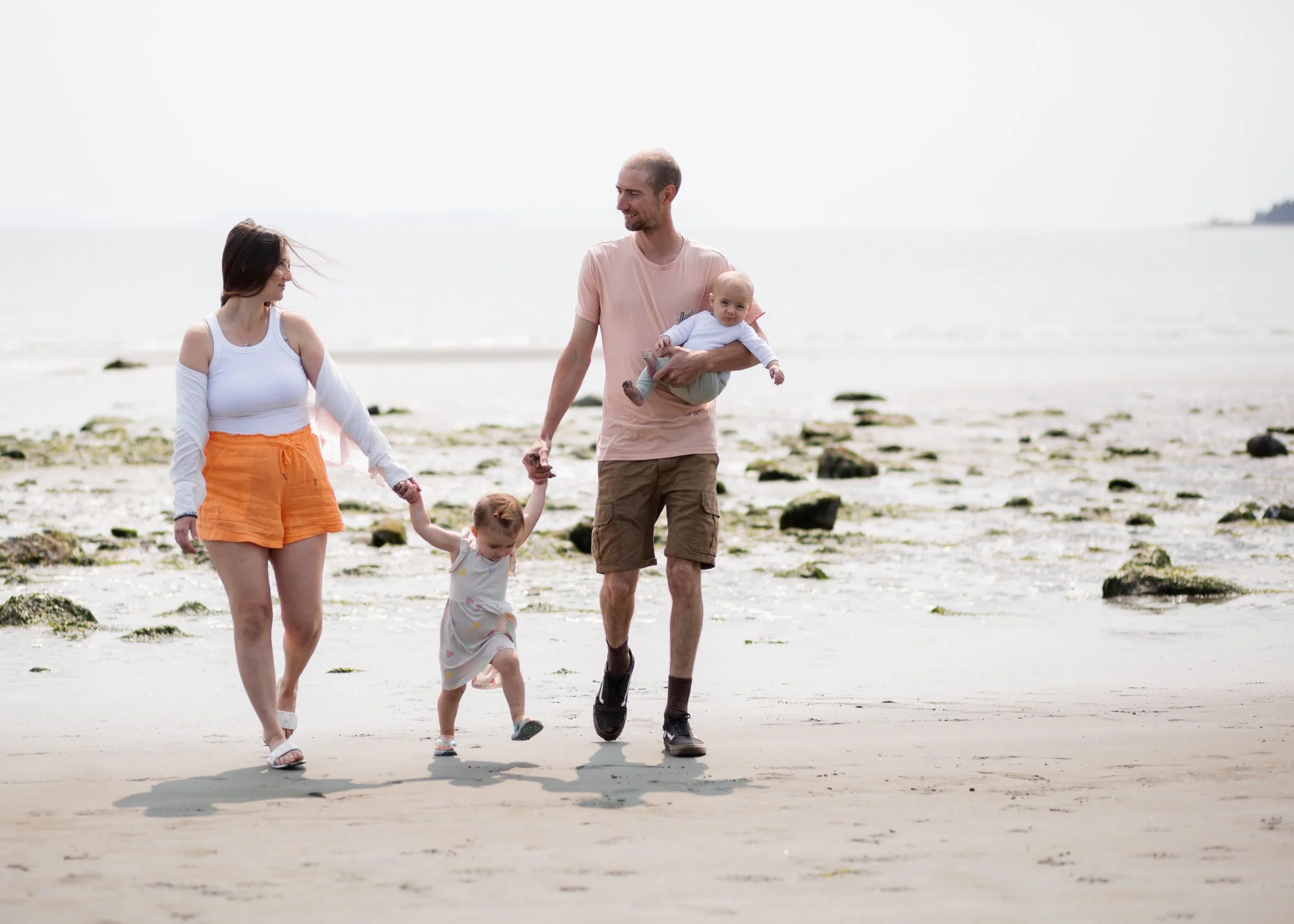 A family walking on a beach, holding hands, with the father carrying a baby and the mother walking alongside two young children.