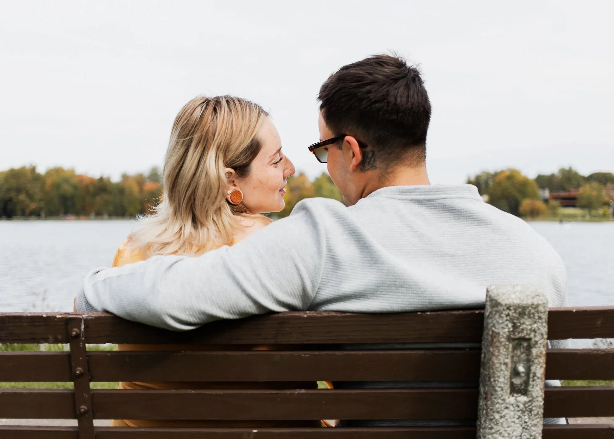 A couple sitting on a bench by a lake, enjoying the outdoors, with fall foliage in the background.