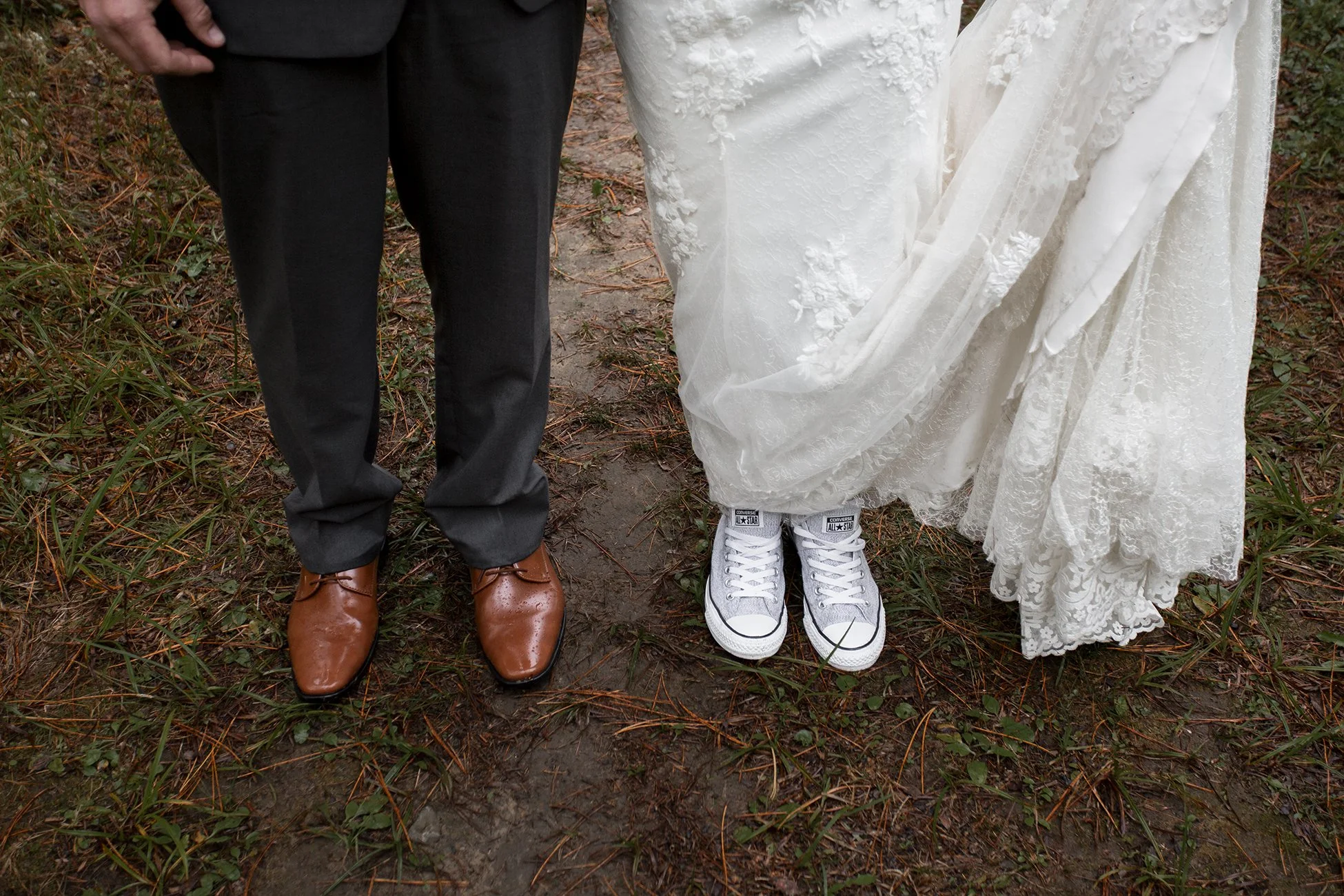 Close-up of a bride and groom's feet standing on a dirt path with grass on the sides, the bride is wearing white lace wedding gown and Converse sneakers, the groom is in brown dress shoes and black pants.