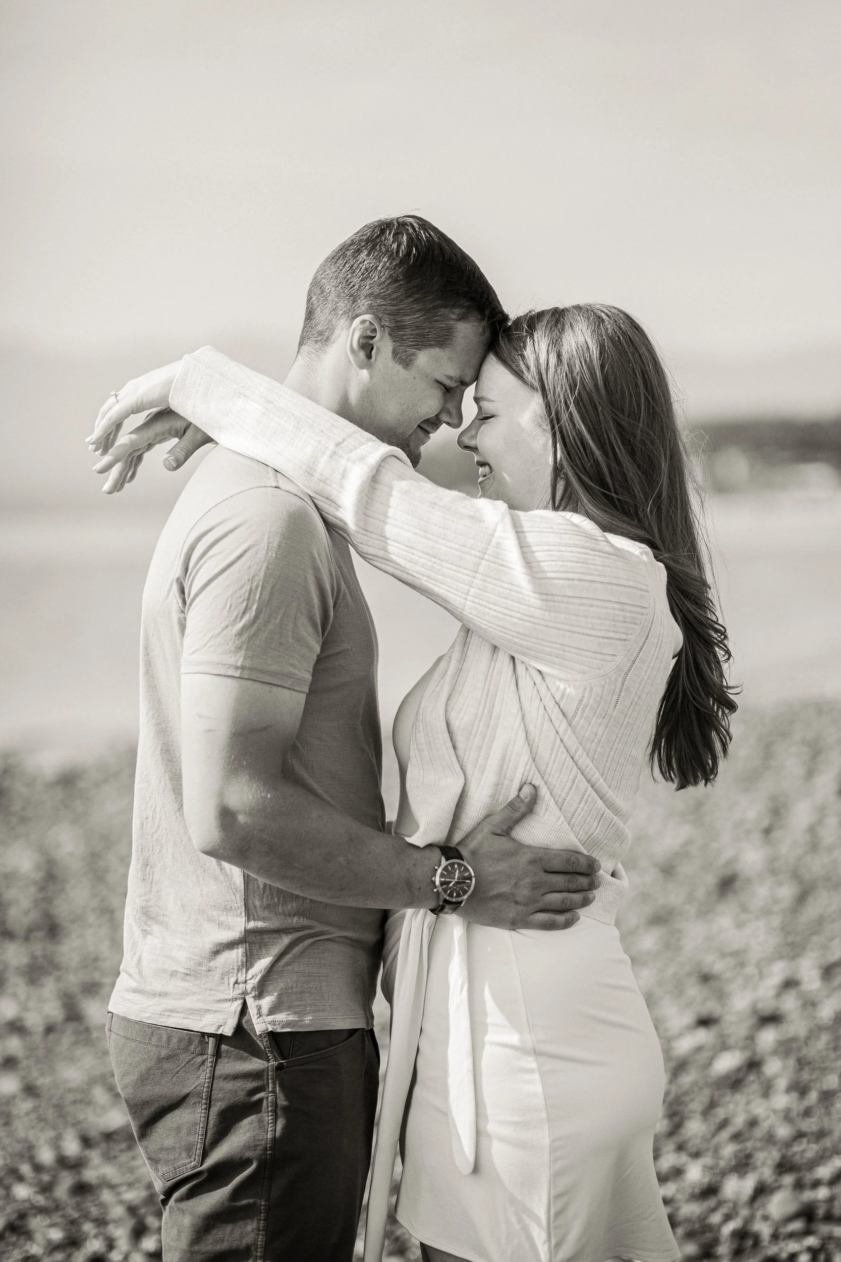 A black and white photo of a couple embracing each other closely on a beach, with their foreheads touching and eyes closed, smiling gently.