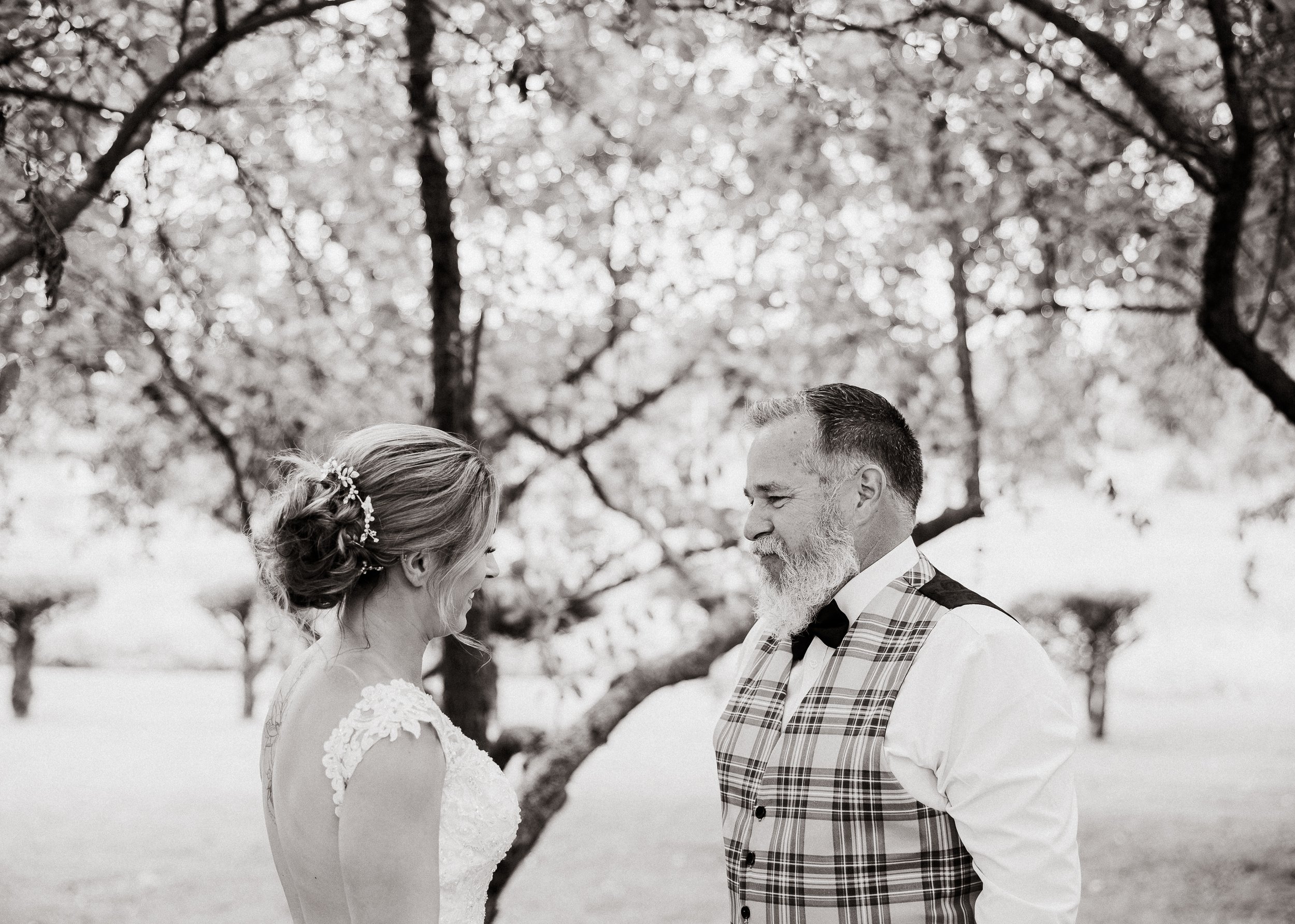 A bride and groom standing close to each other outdoors, holding hands and smiling, with trees in the background.