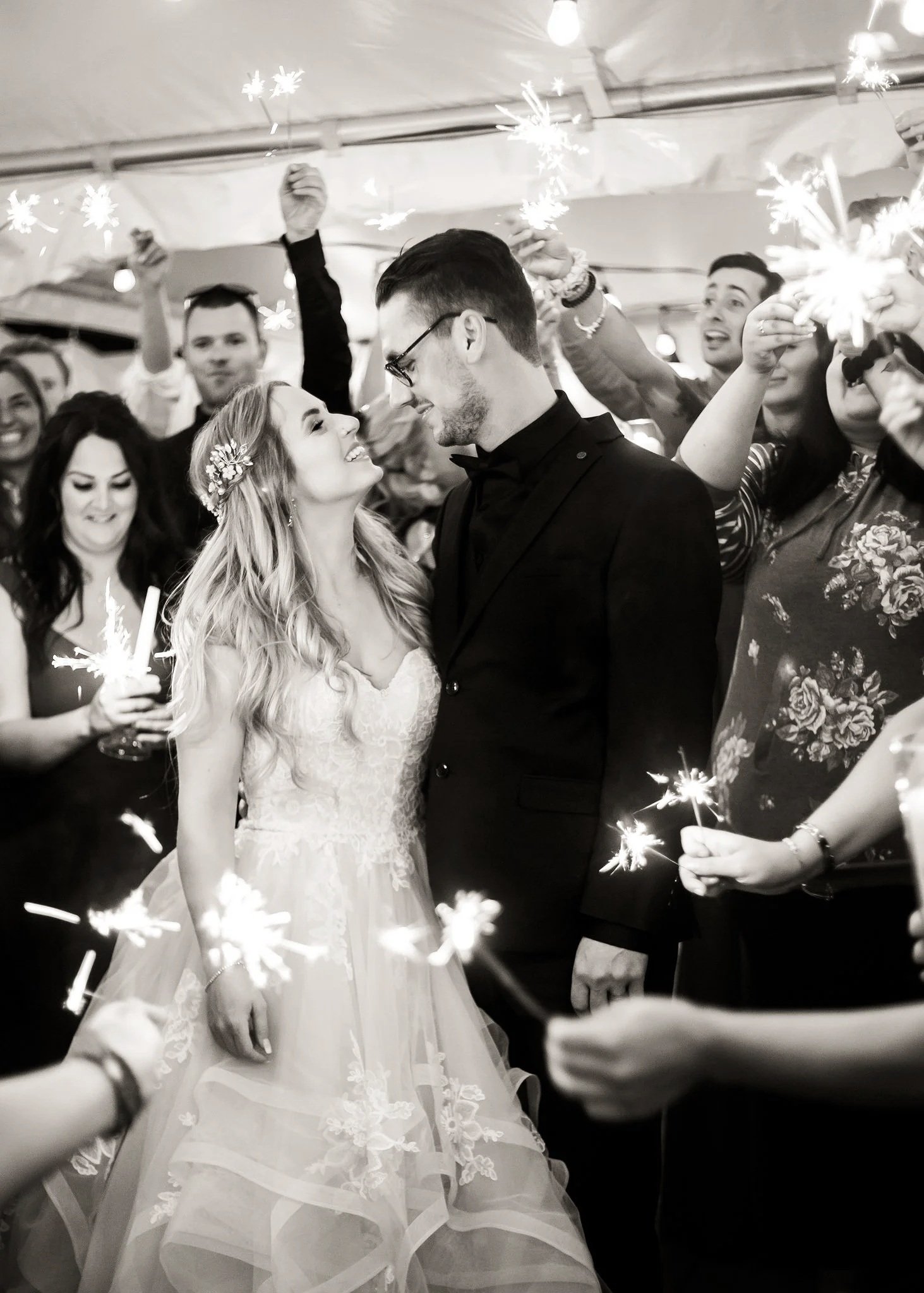 A wedding couple, the bride and groom, are smiling and looking at each other, surrounded by guests holding sparklers at a celebration.
