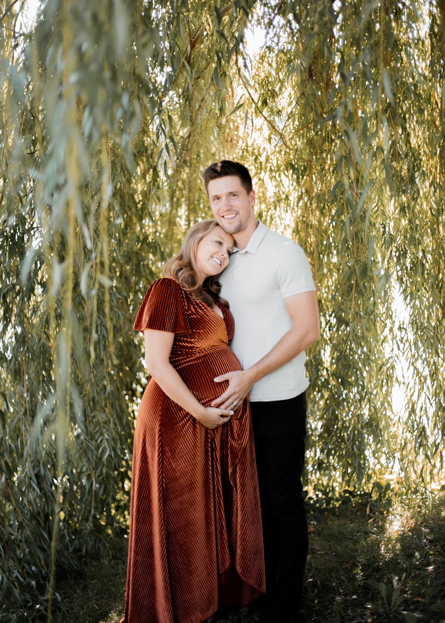 A smiling pregnant woman and a man are standing close together, with the woman resting her head on the man's shoulder. The woman is wearing a rust-colored, velvet, pleated dress, and the man is dressed in a white polo shirt and black pants. They are outdoors, surrounded by hanging green leaves of a tree, with sunlight filtering through the foliage, creating a warm, golden glow.