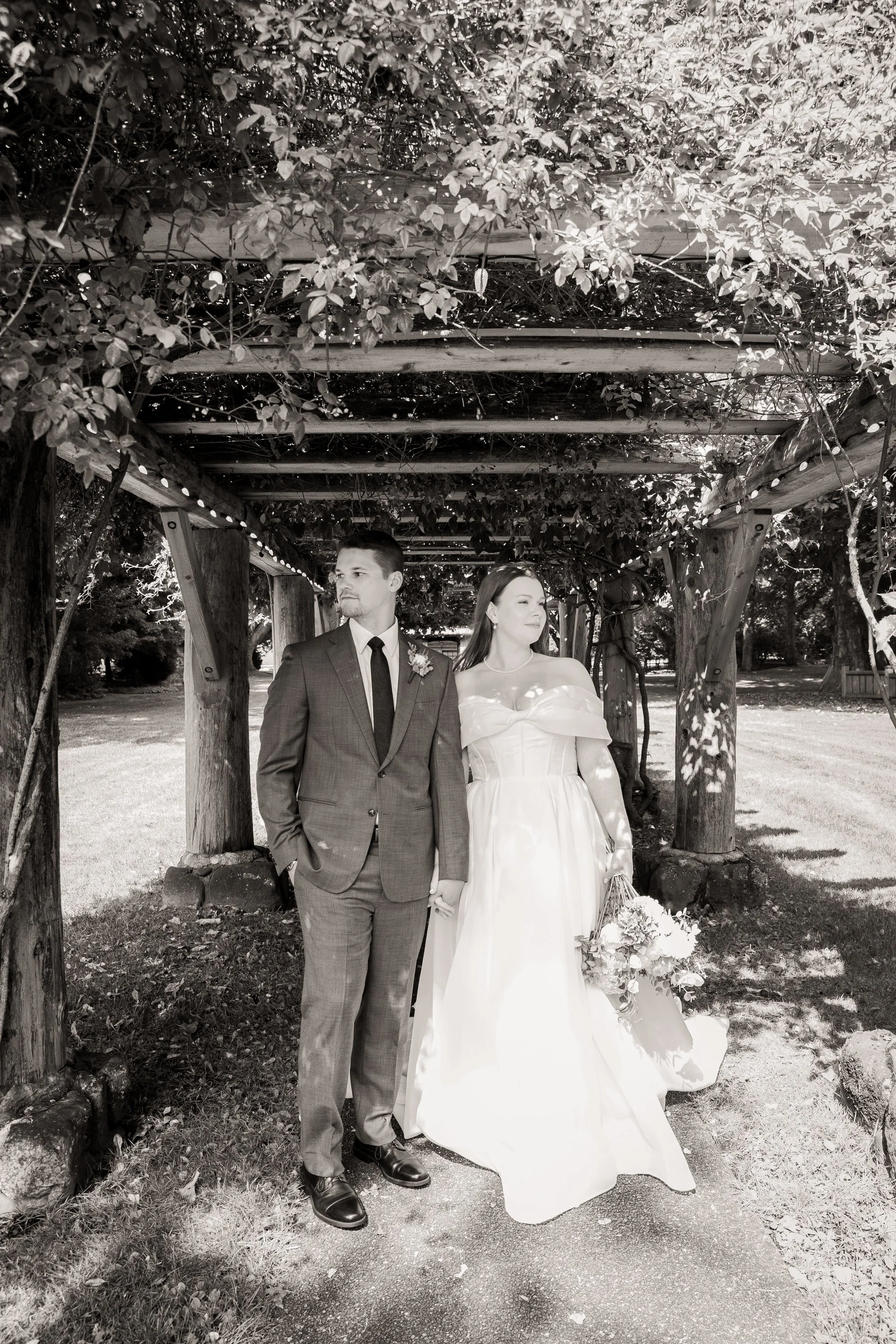 A black and white photo of a bride and groom standing hand in hand under a wooden arbor with leafy vines. The groom is wearing a suit and tie, and the bride is in a strapless wedding gown holding a bouquet. They are outdoors on a sunny day with trees and grass in the background.