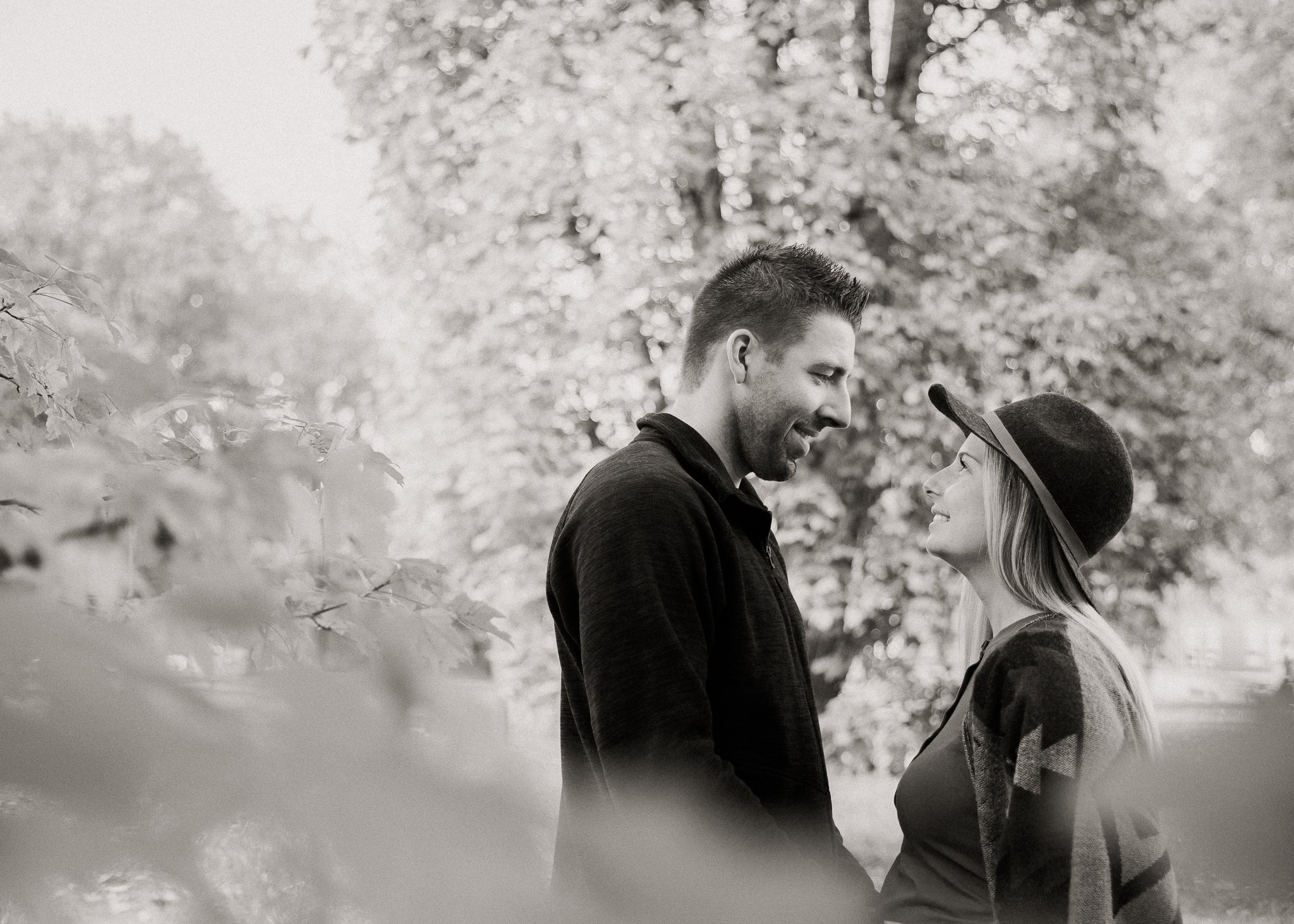 A black and white photo of a couple standing outdoors in nature, looking at each other and smiling softly.