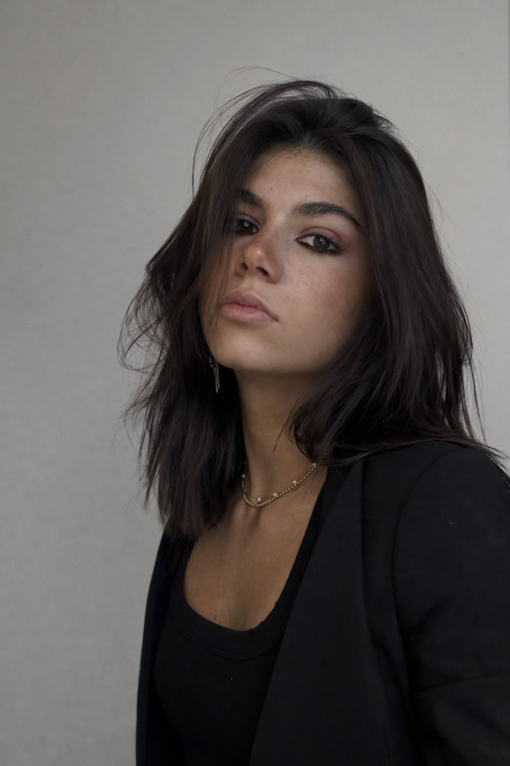 A woman with shoulder-length dark hair and light skin, wearing a black top, a black blazer, and gold jewelry, looking at the camera with a neutral expression against a plain background.