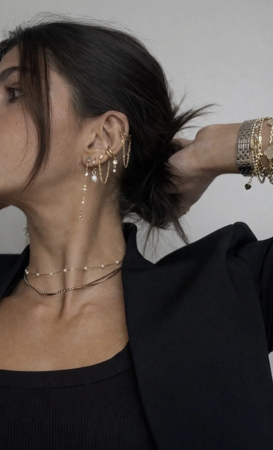 Close-up of a woman's left profile showing her ear with multiple gold earrings, her neck with layered necklaces, and her wrist with various gold and silver bracelets. She is wearing a black top and a black blazer, with her hand behind her head, revealing dark brown hair.