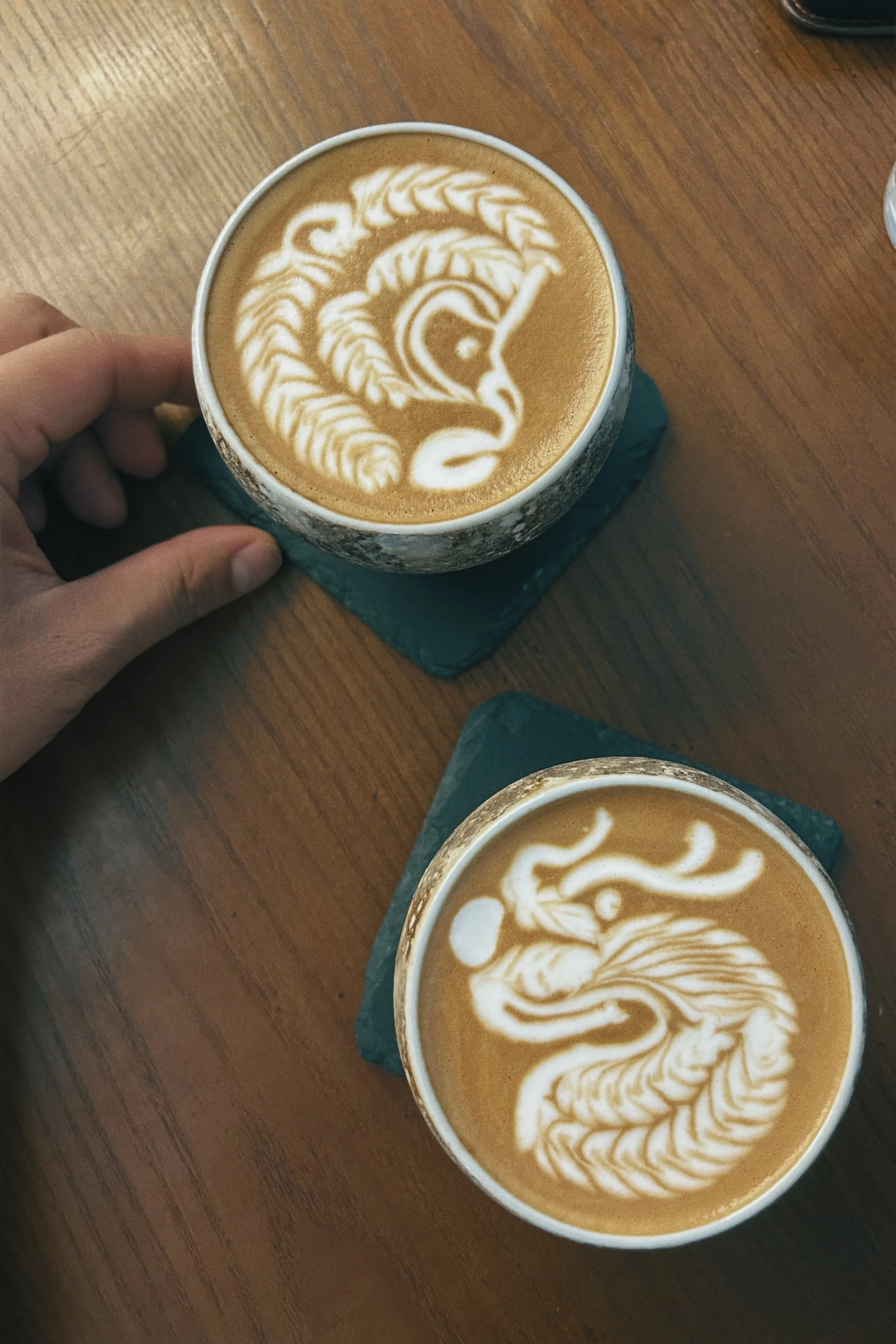 Two cups of coffee with latte art swans on top, placed on green coasters on a wooden table. One hand is holding the cup on the left side of the image.