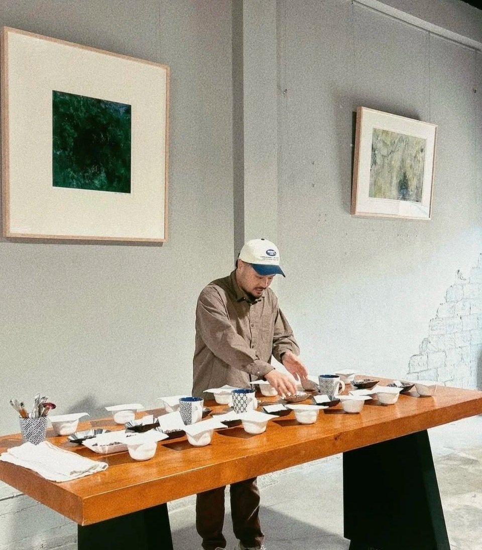 A man wearing a beige shirt, dark pants, and a white cap is arranging dishes and cups on a large wooden table in a room with gray walls, two framed pictures hanging on the wall, and a partially exposed brick wall on the right side.