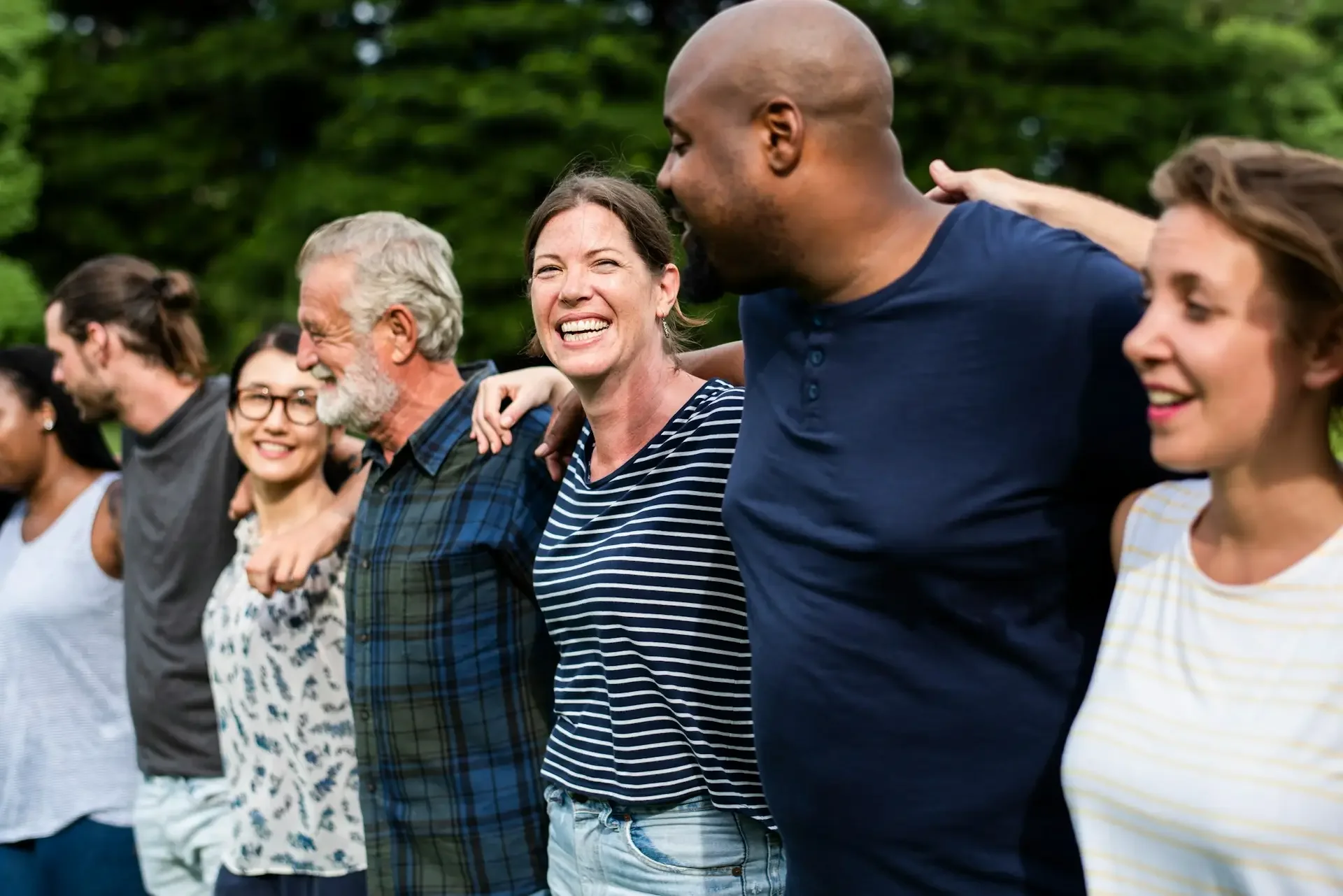 Group of people standing outdoors with their arms around each other's shoulders, smiling and laughing.