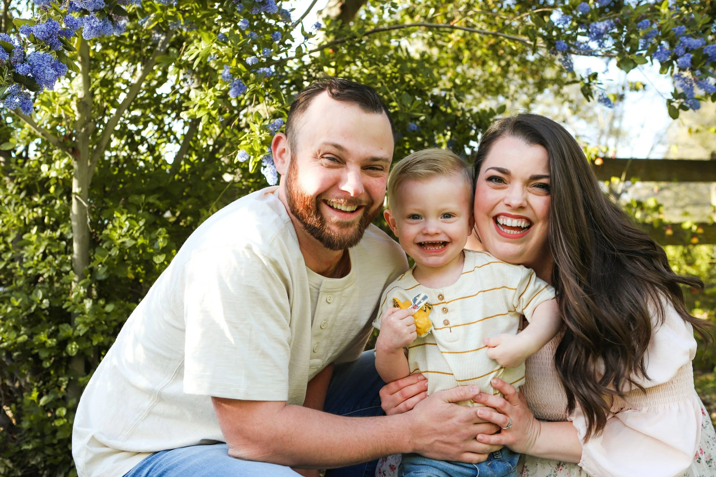 A smiling family of three with a man, woman, and young girl outdoors, standing in front of green bushes with purple flowers, enjoying sunny weather.
