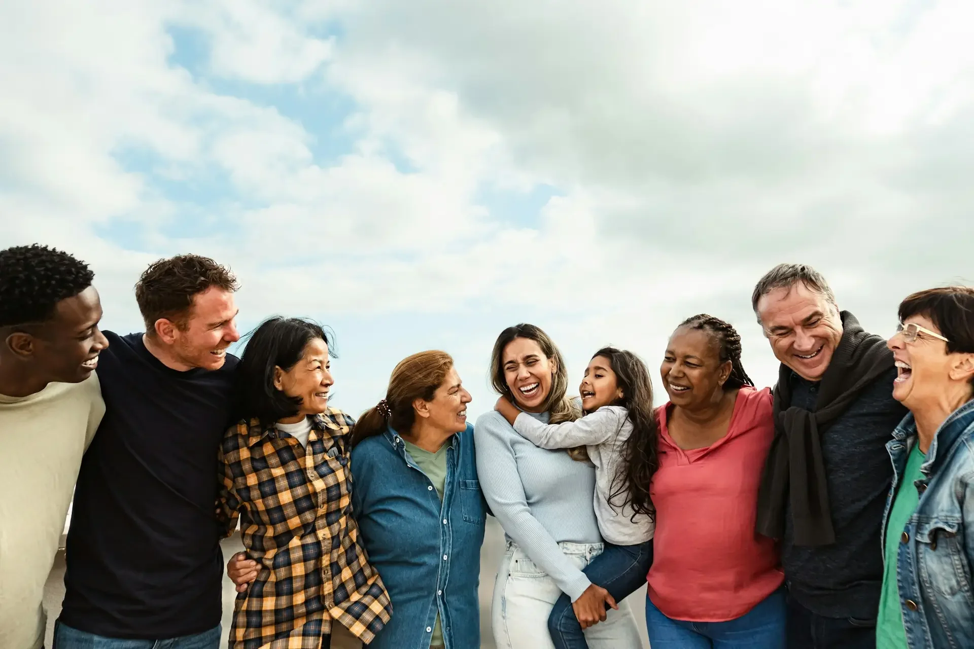 A diverse group of people standing outdoors in a circle, laughing and smiling, under a partly cloudy sky.