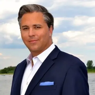 A man in a dark suit with a light blue pocket square standing outdoors near a body of water with a cloudy sky in the background.