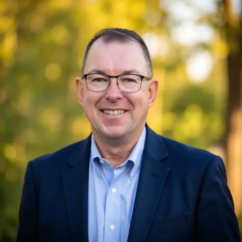 A smiling man in glasses wearing a blue suit and light blue collared shirt outdoors with blurred green trees in the background.