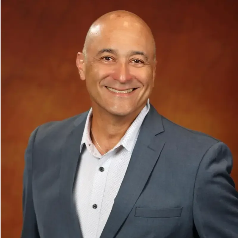 A smiling man in a gray suit and white shirt standing against a brown background.