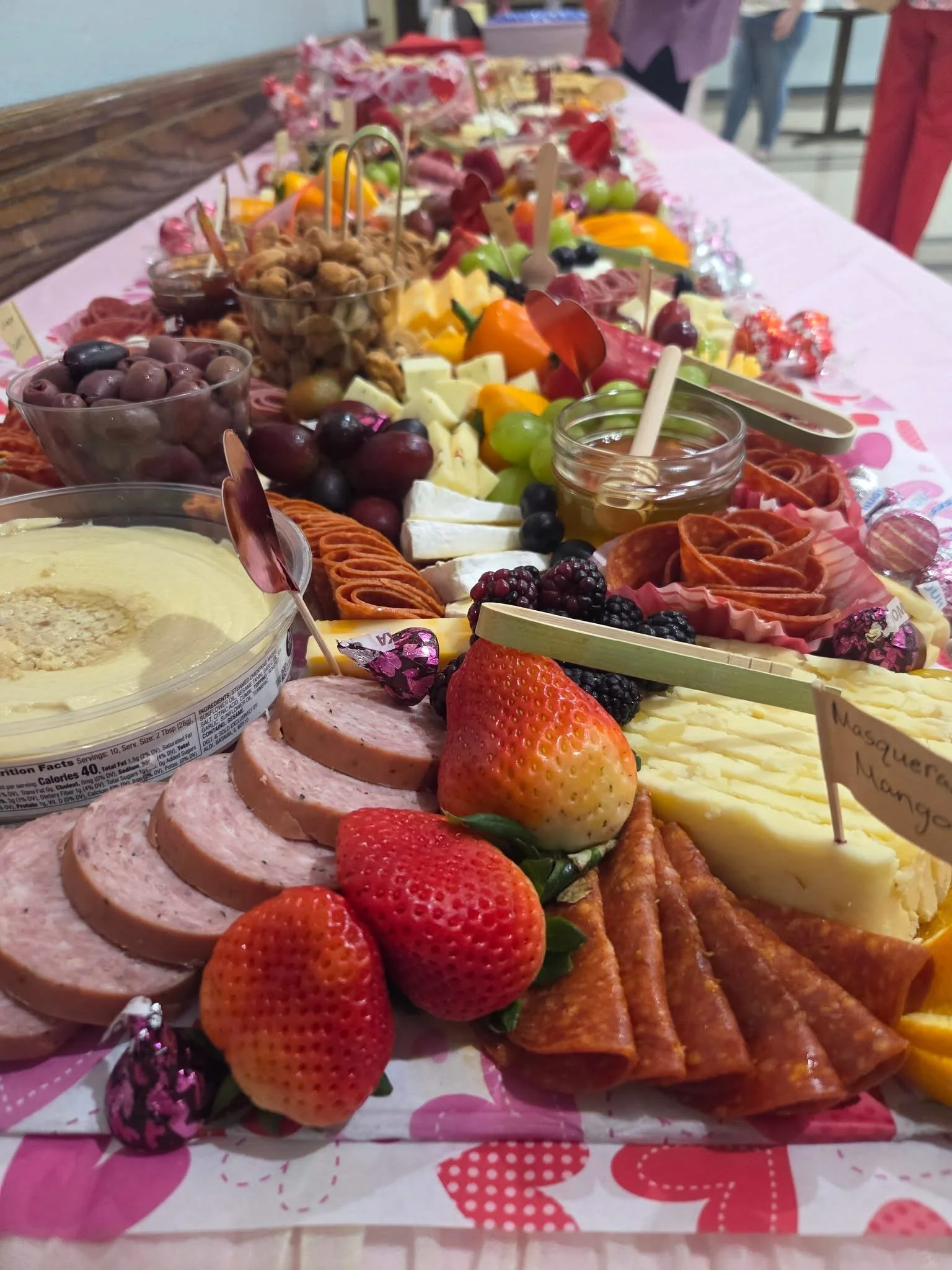 A large breakfast or brunch spread on a table, featuring sliced strawberries, sliced sausage, cheese, grapes, blackberries, chocolate candies, honey, sliced meats, and various other small food items.