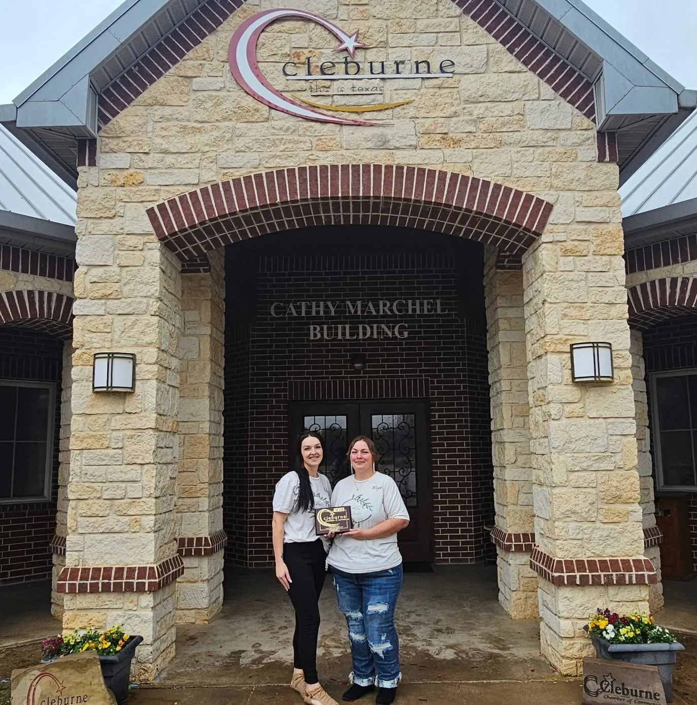 Two women standing in front of the Cleburne Chamber of Commerce building, holding an award or plaque, smiling for the photo.