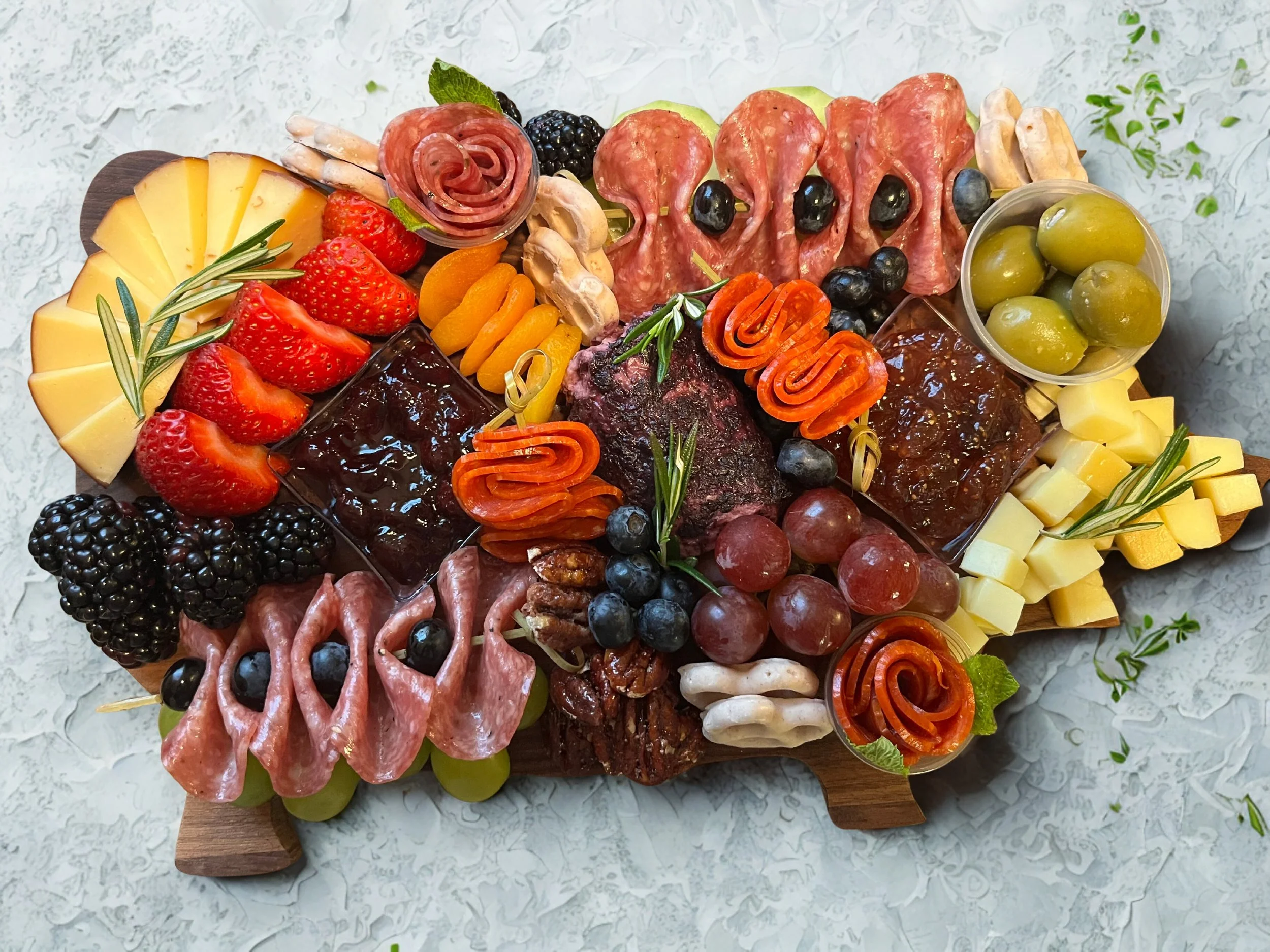 Assorted fruit and charcuterie platter with cheese, grapes, strawberries, blackberries, blueberries, green olives, sliced meats, jam, and garnishes on a wooden serving board.