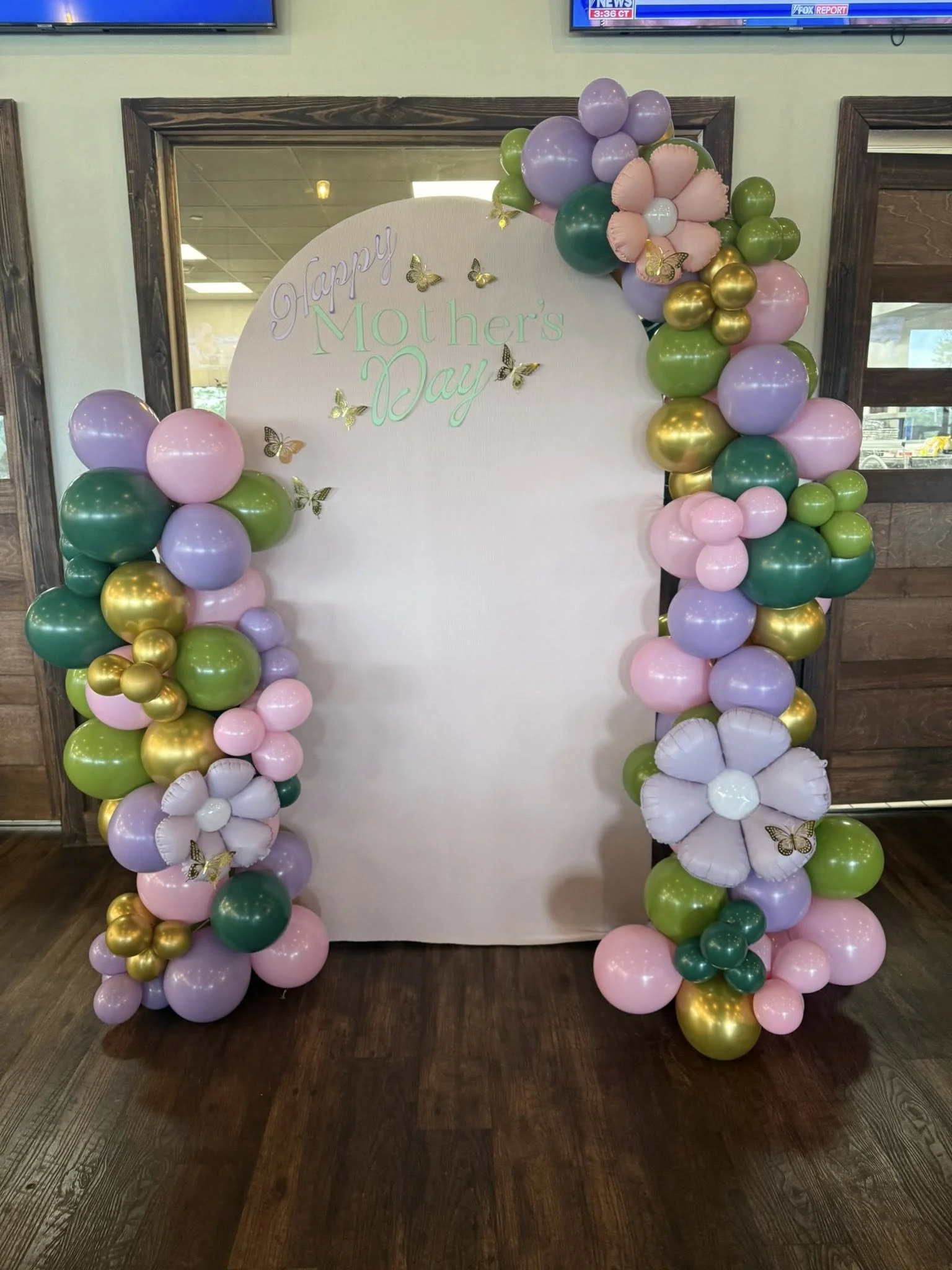 A Mother's Day backdrop decorated with colorful balloons in shades of pink, purple, green, gold, and white, with butterfly and flower embellishments, displaying the message "Happy Mother's Day."