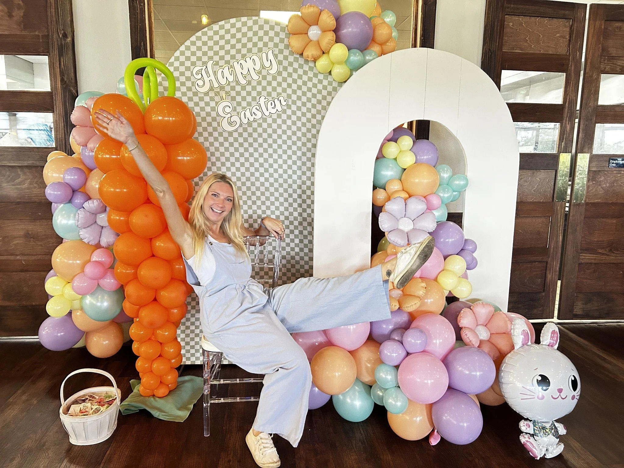 Woman smiling, sitting on a transparent acrylic chair, with one leg raised, surrounded by colorful pastel balloons and a cheerful Easter-themed backdrop that says 'Happy Easter'.