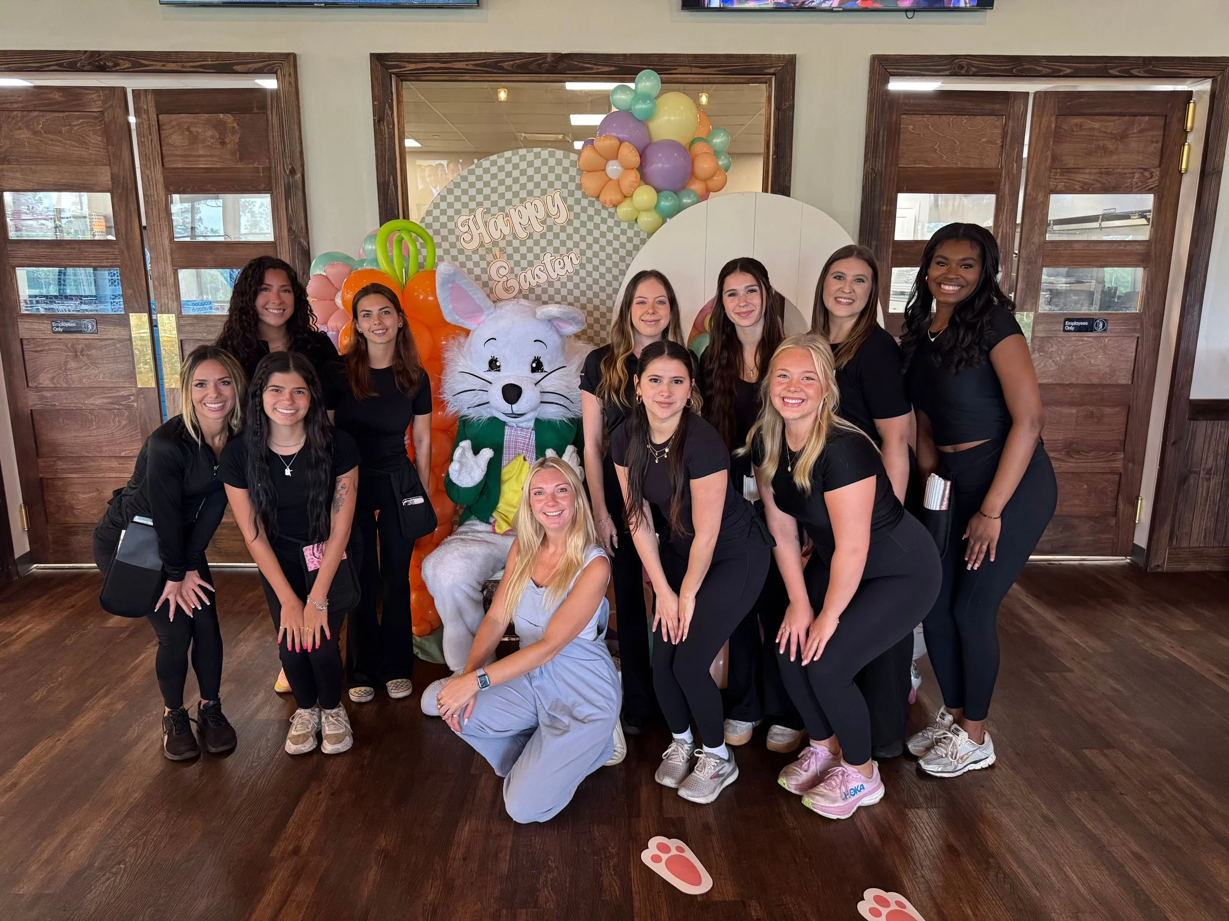 Group of 11 women posing with an Easter bunny mascot in front of Easter-themed decorations, including balloons and a 