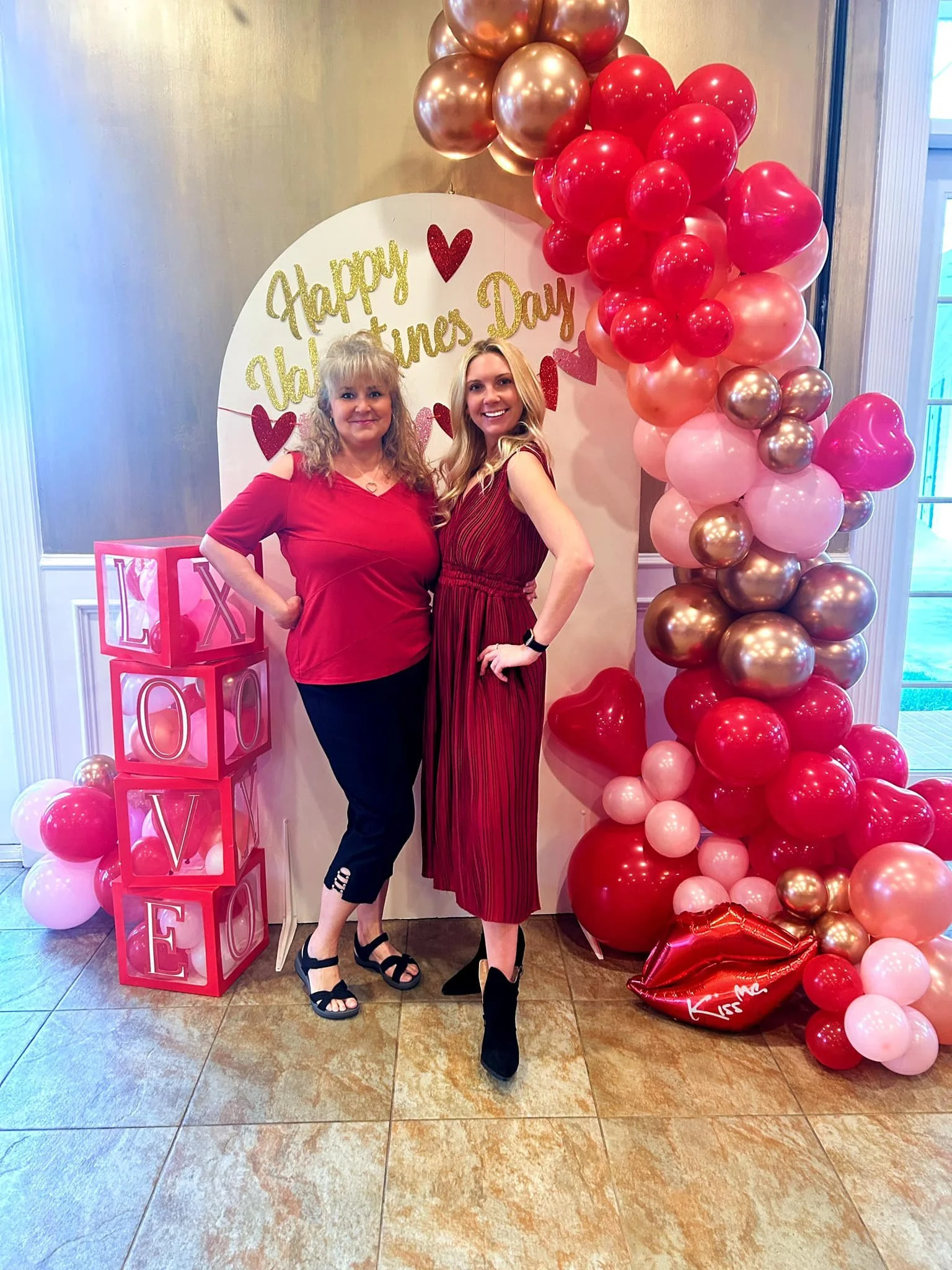 Two women standing in front of a Valentine's Day and love-themed background with pink, red, and gold balloons, a sign that says 'Happy Valentine's Day,' and a pink block display with the letters 'L O V E' on it.