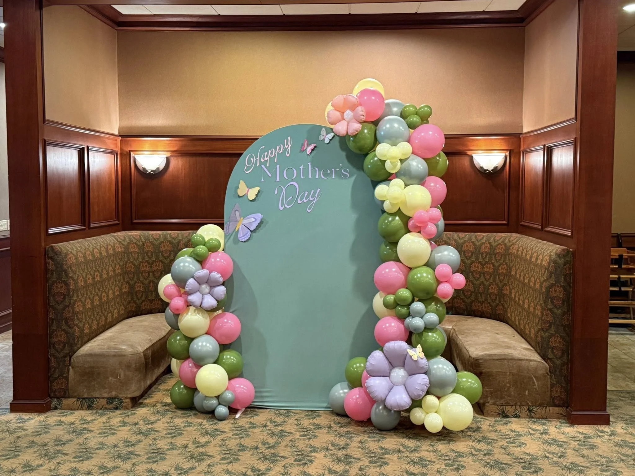 Balloon arch with pink, yellow, green, and light blue balloons, and butterfly decorations, set up for Mother's Day on a teal backdrop with the words 'Happy Mother's Day' in a festive font.
